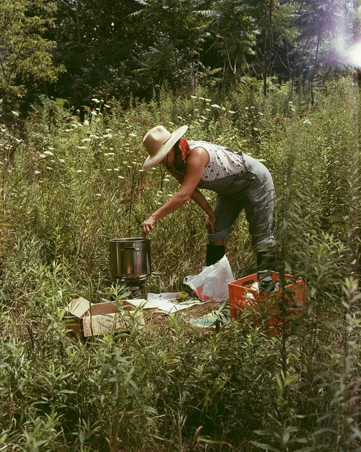 In a densely vegetated field of tall grass and wildflowers, a person in overalls bends over a pot, mixing it. They are surrounded by other miscellaneous supplies, boxes, and crates.