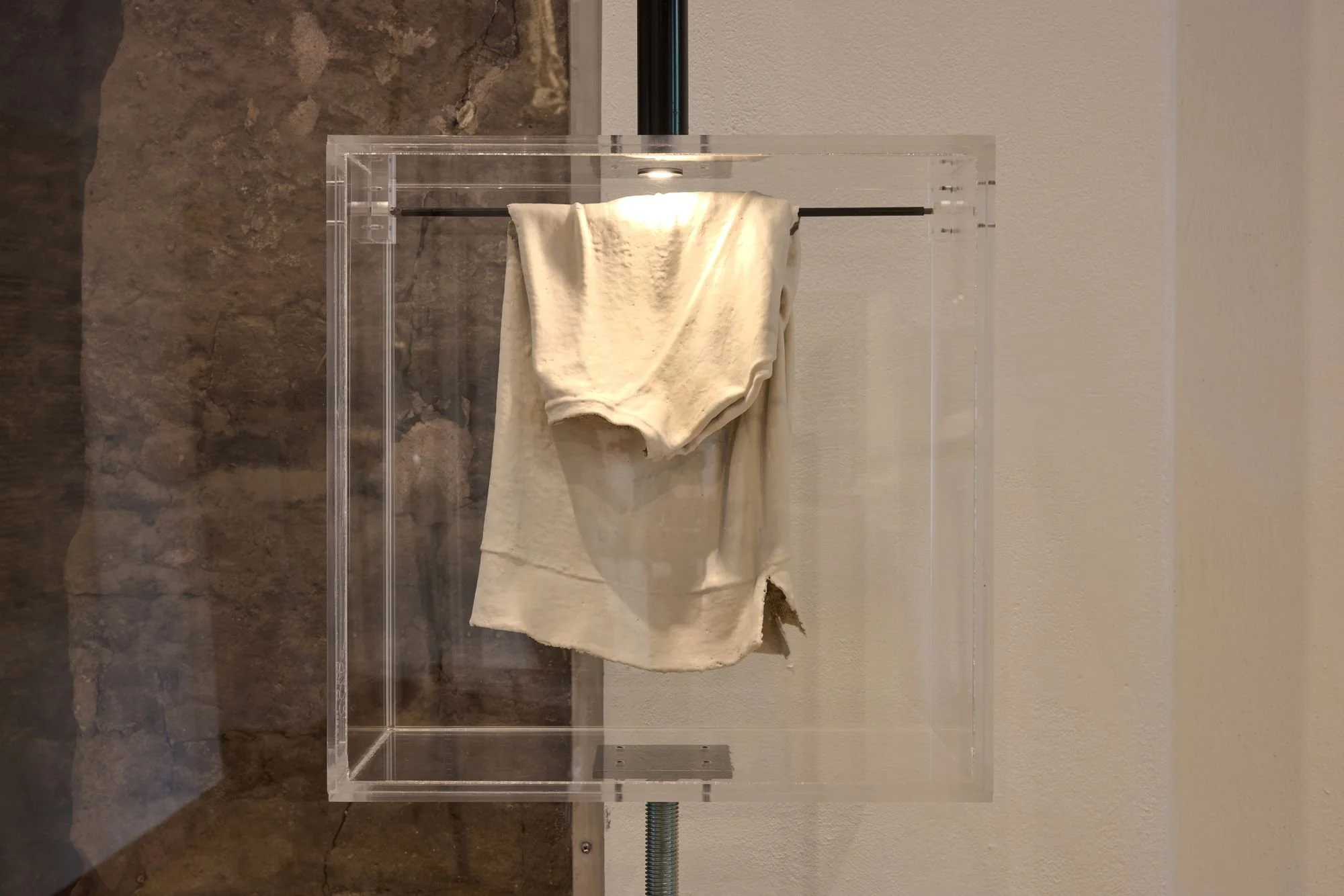 Worn white garment suspended inside a clear acrylic display case mounted on a metal pole.
