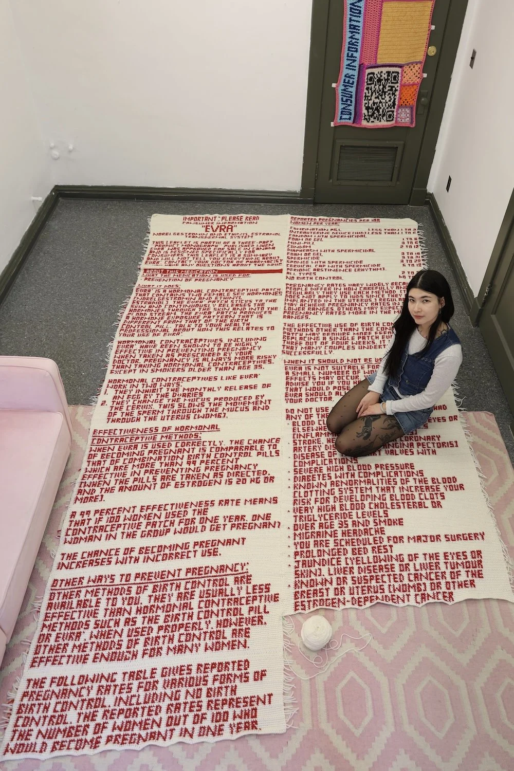 Artist sitting on the floor atop a crocheted red-text textile installation, surrounded by additional textile works hanging on the walls.