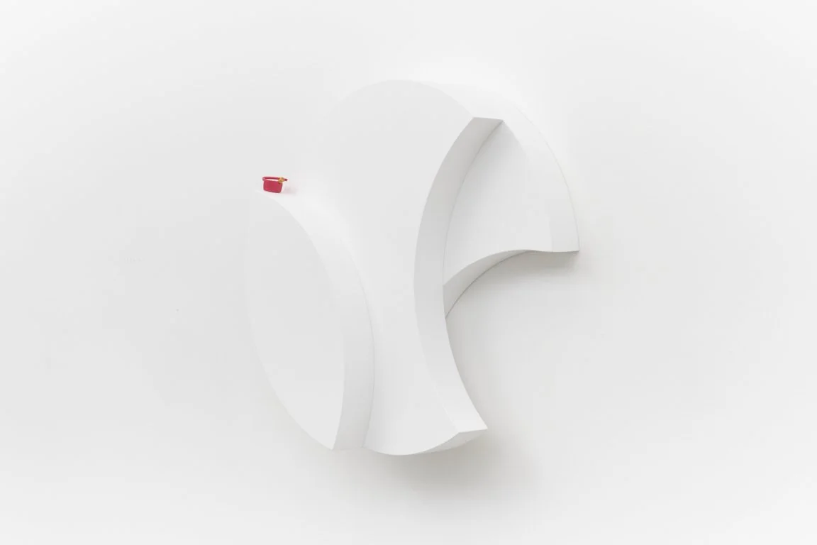 A white sculpture mounted to a white wall consists of overlapping rounded shapes with a small red bottle cap resting on the top of one of the forms.