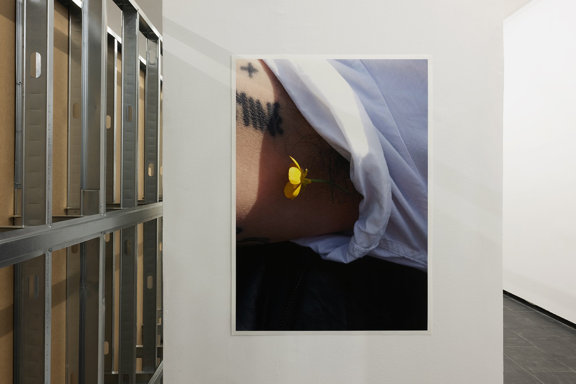 Photograph of a person’s torso partially covered by white fabric, with a small yellow flower resting against the skin near a tattoo.