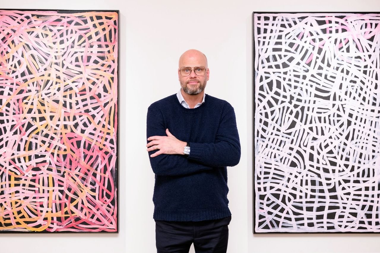 A man in a blue sweater with glasses and a beard stands in between two paintings of the same size and style. The paintings feature curling lines that intersect, almost like a street map. One painting has a pink and yellow palette, the other is white.