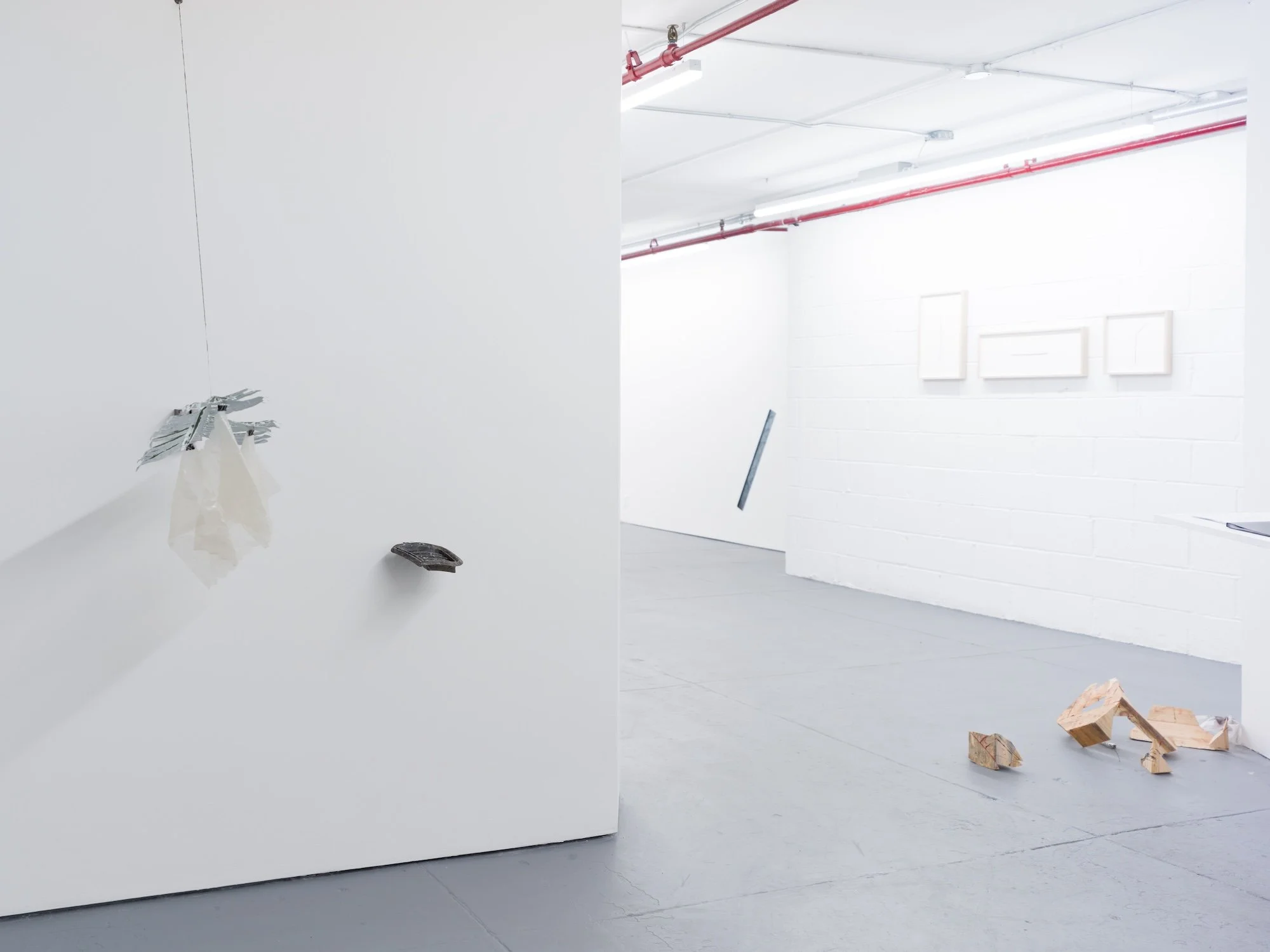 Gallery installation view with hanging translucent sculpture, wall-mounted piece, and scattered wooden fragments on the floor.