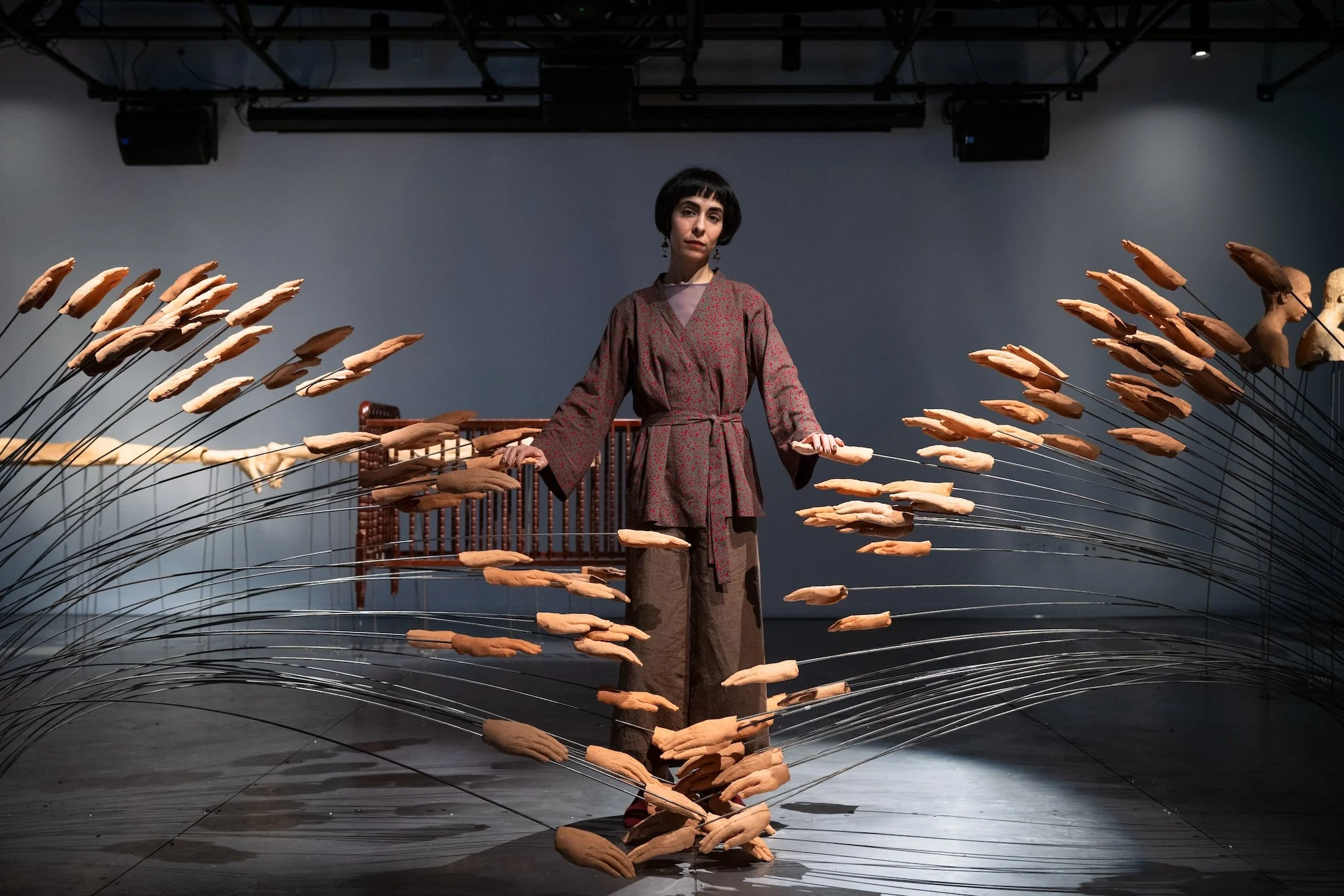 In a large gallery, a person in brown, flowing clothes rests their hands on a sculpture, which is comprised of multiple hands affixed to flexible metal rods stemming from two sides with the hand ends meeting in the middle.
