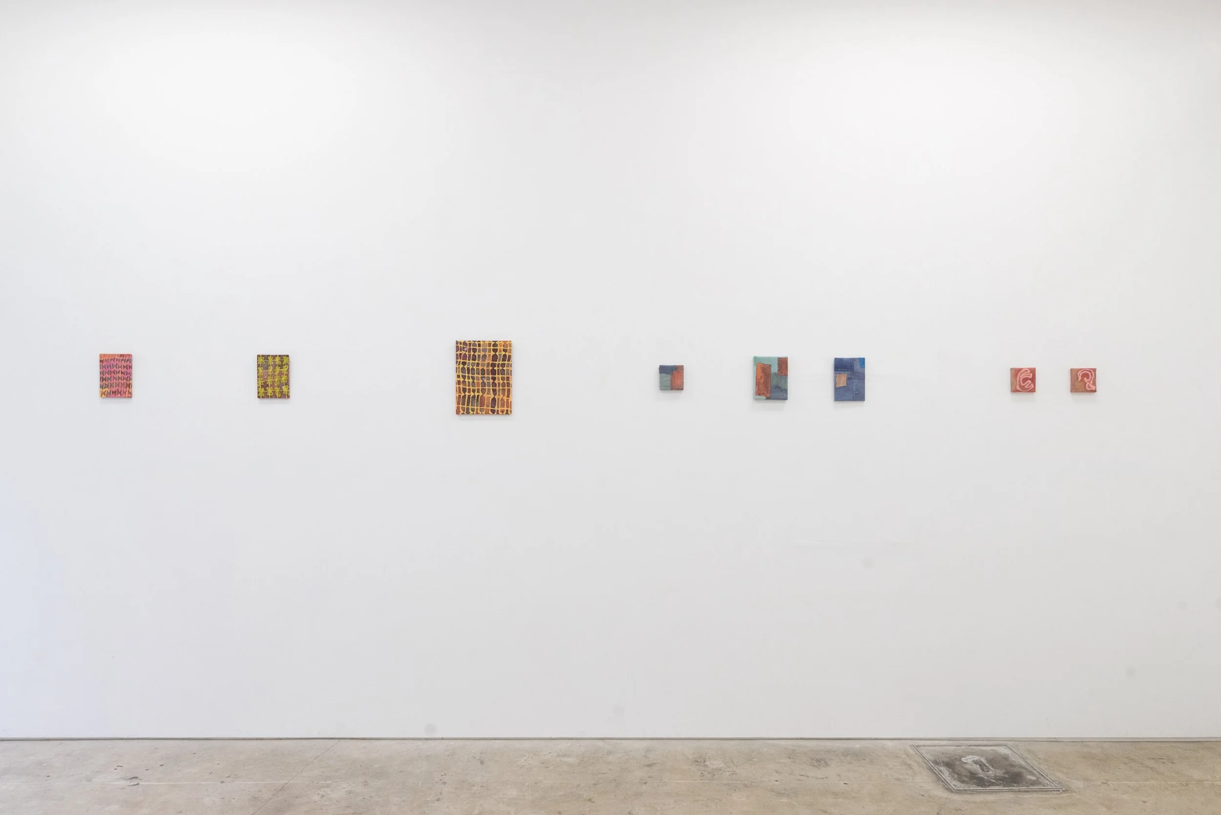 A white gallery wall features a number of small, rectangular works. Each features soft colors and cross-weave patterns.