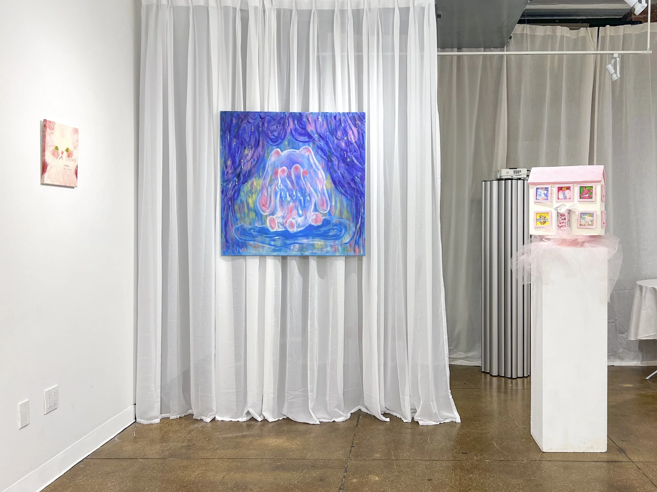 Exhibition space with a sheer white curtain backdrop, a blue abstract painting centered, and a small pink house sculpture displayed on a pedestal to the right.
