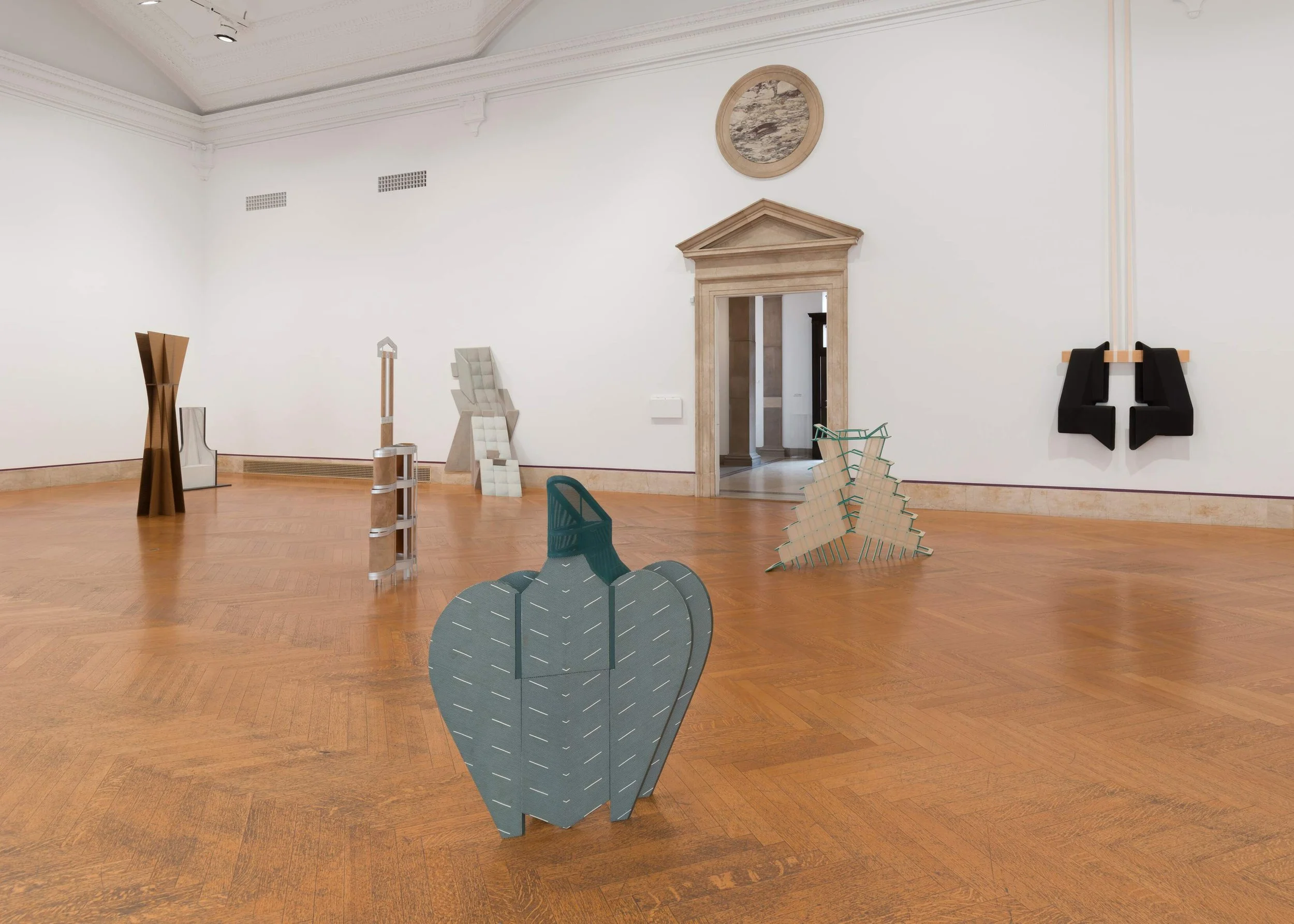 A white-walled room with hardwood floors features a number of wall mounted and free-standing sculptures. Each sculpture is visually distinct, but all are structural and geometric and of medium size.