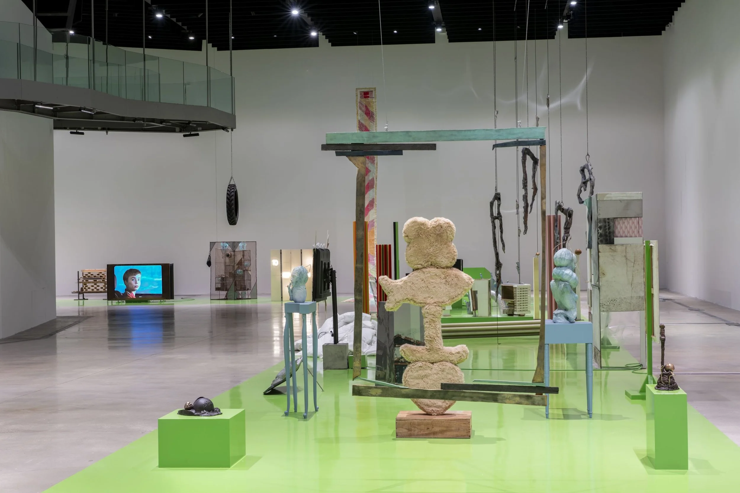 A large, white gallery room with a second-story glass balcony has a strip of green flooring running centrally throughout the room. On the green strip is a collection of sculptures, all visually distinct, but grouped closely together.