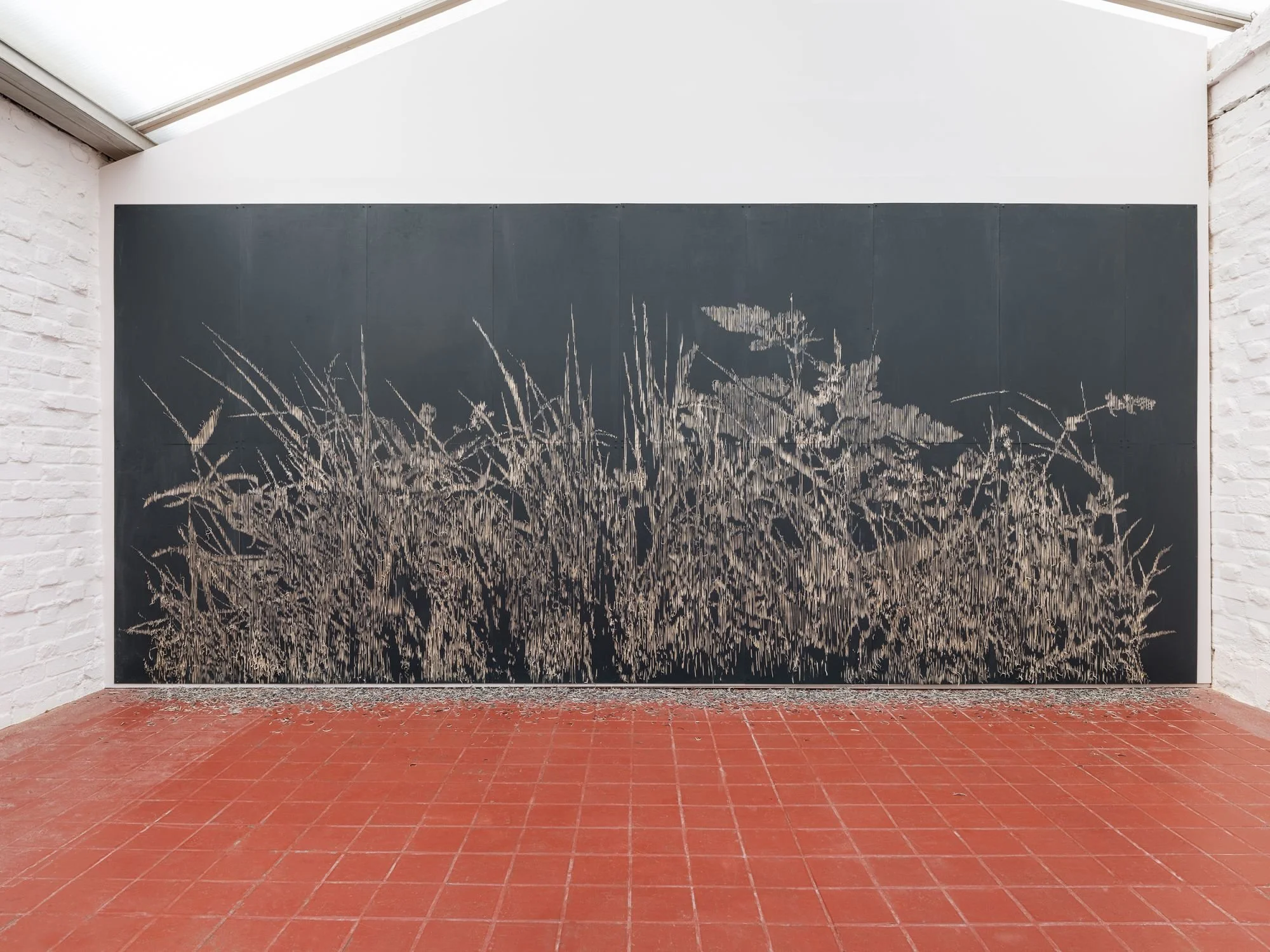 A black wall within a white structure bears marks that resemble a line of long grass or wheat. The terracotta tiled floor bears residue that resembles the grass.