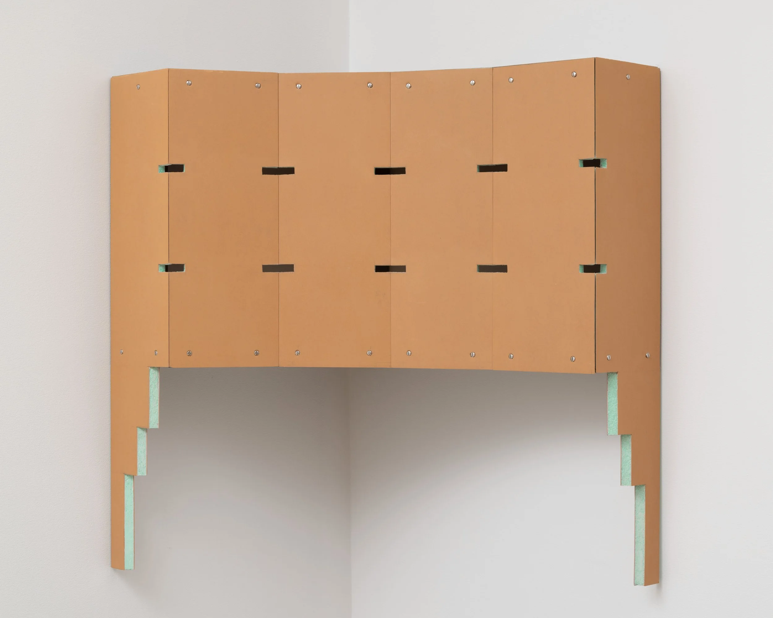 A sculpture, seemingly made from wooden board or cardstock, features pleats and folds, mounted to the corner of two walls. The sculpture also features evenly equidistant small rectangular holes. Each side has a stair-like descending bottom edge.