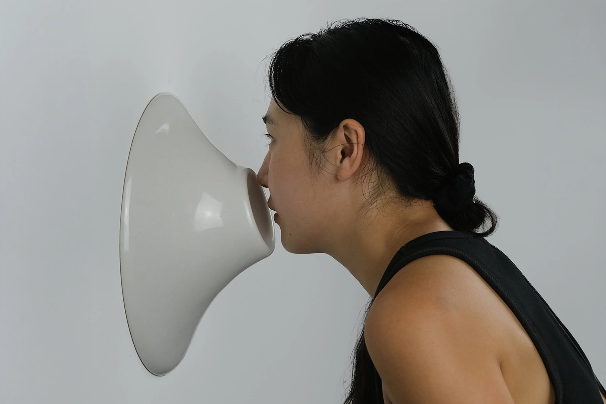 A woman in profile presses her face into a smooth, white, wall-mounted sculptural form against a plain background.