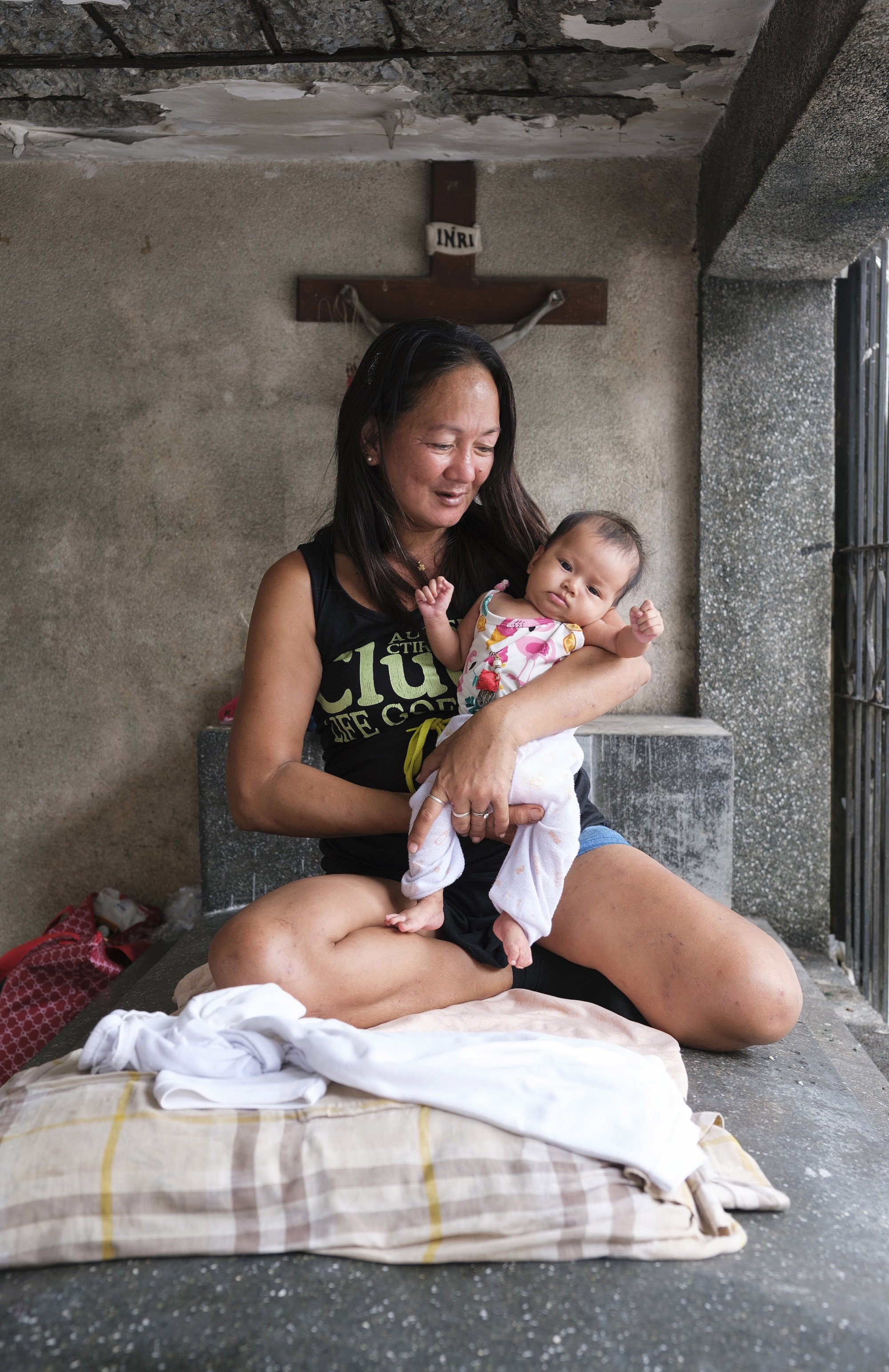 Woman sitting inside a concrete tomb holding a baby, with a crucifix mounted on the wall behind them.