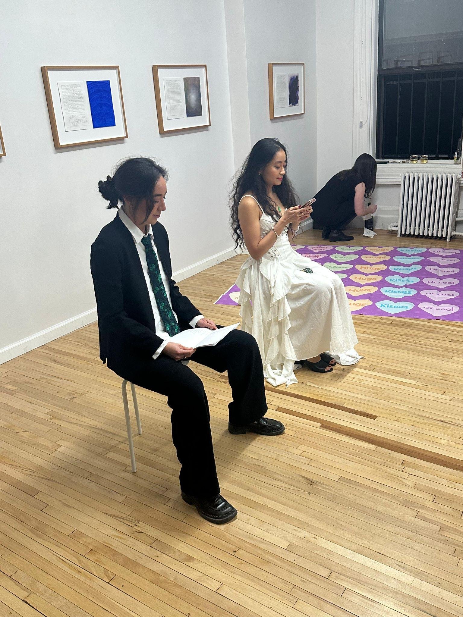 Two people sit on stools in an art gallery; one wears a black suit and tie while reading, and the other wears a white ruffled dress while looking at a phone.