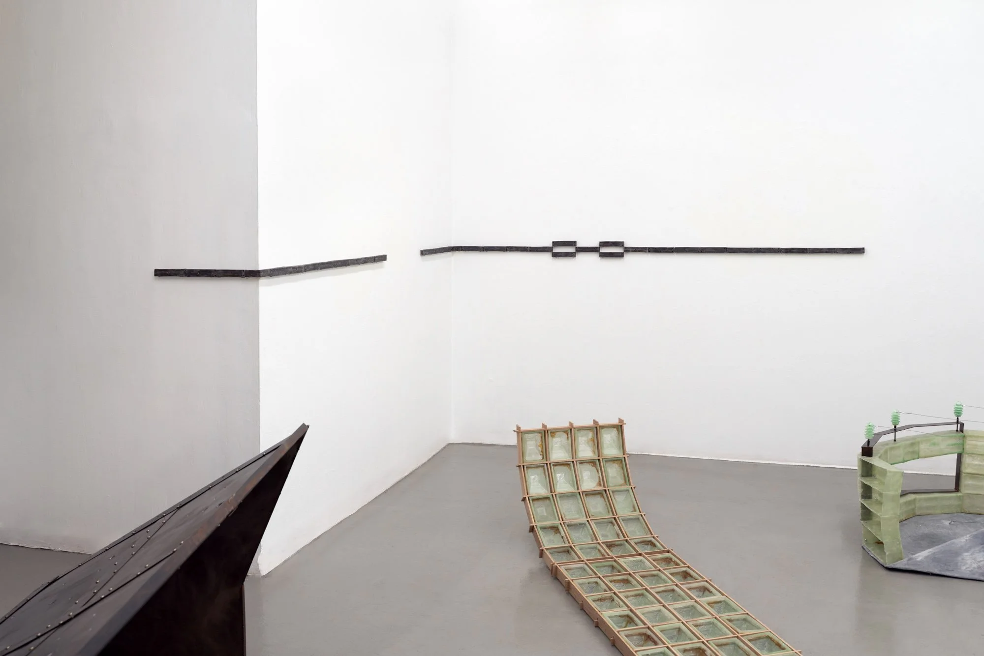 Corner of a white gallery with a narrow wall-mounted metal strip and floor sculptures in green resin and steel.
