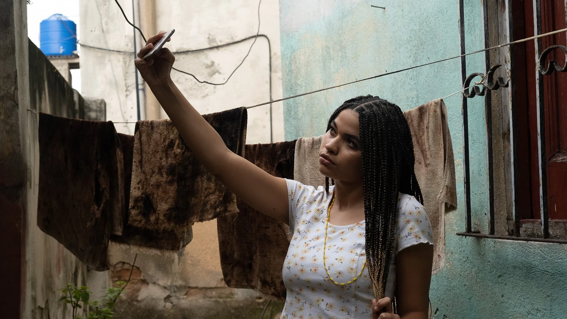 A girl with long box raids and a white floral shirt casually twists her hair in one hand while posing for a selfie. Behind her, run-down buildings support a clothesline of worn fabrics between them.