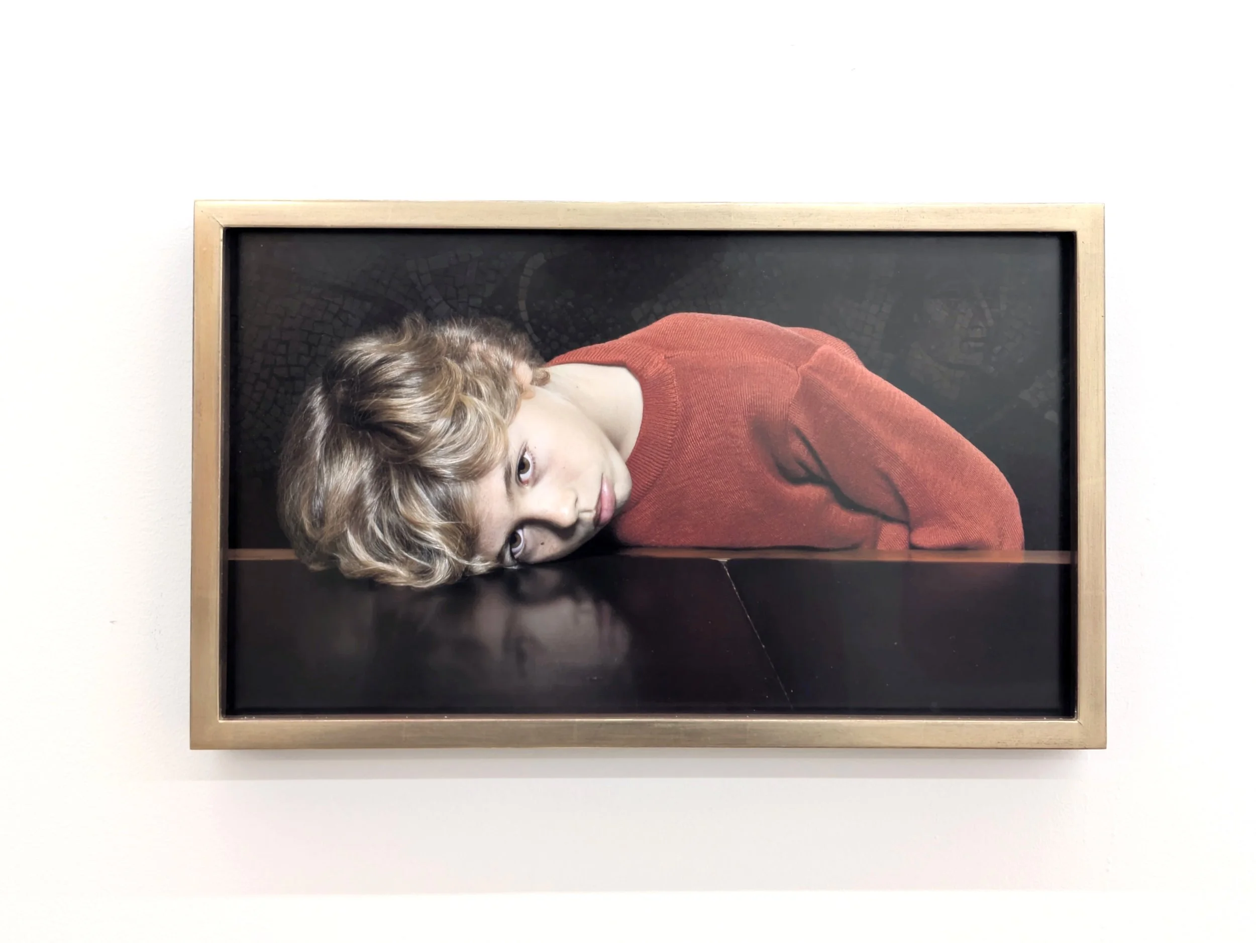 A wall-mounted painting in a plain gold frame features a youthful, blonde figure in a red sweater resting their head on a reflective, black surface. They stare aimlessly into the distance.