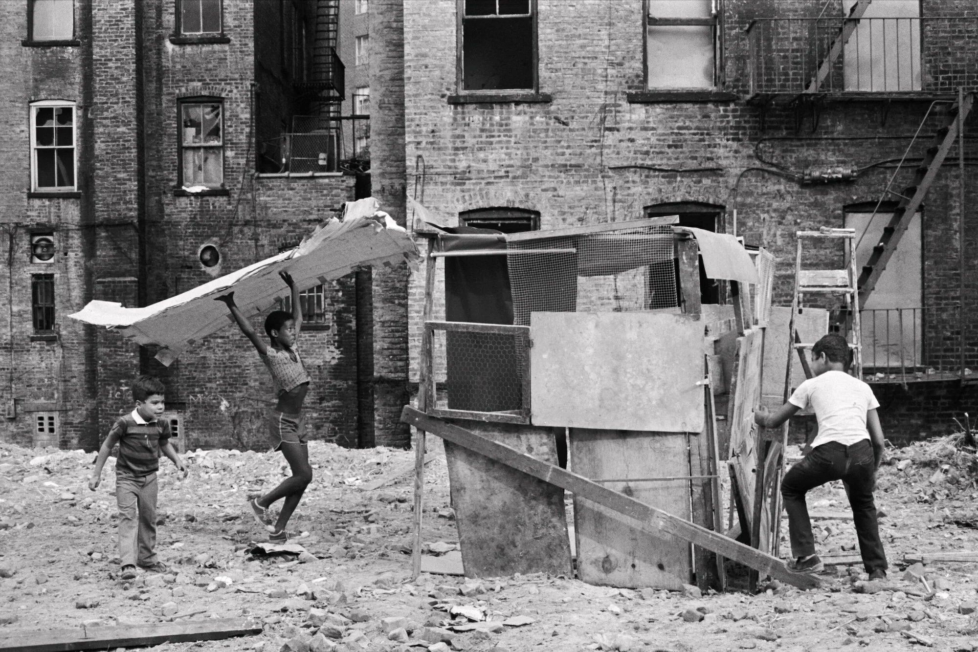 Children build and climb a wooden structure in a rubble-filled vacant lot.