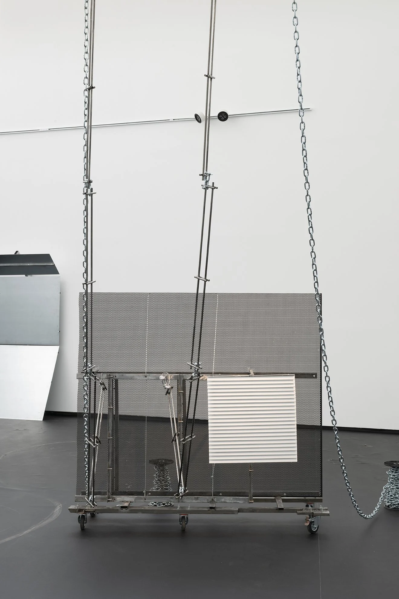 Metal sculpture on wheels with mesh panel, chains, rods, and a white corrugated panel suspended in a gallery space.