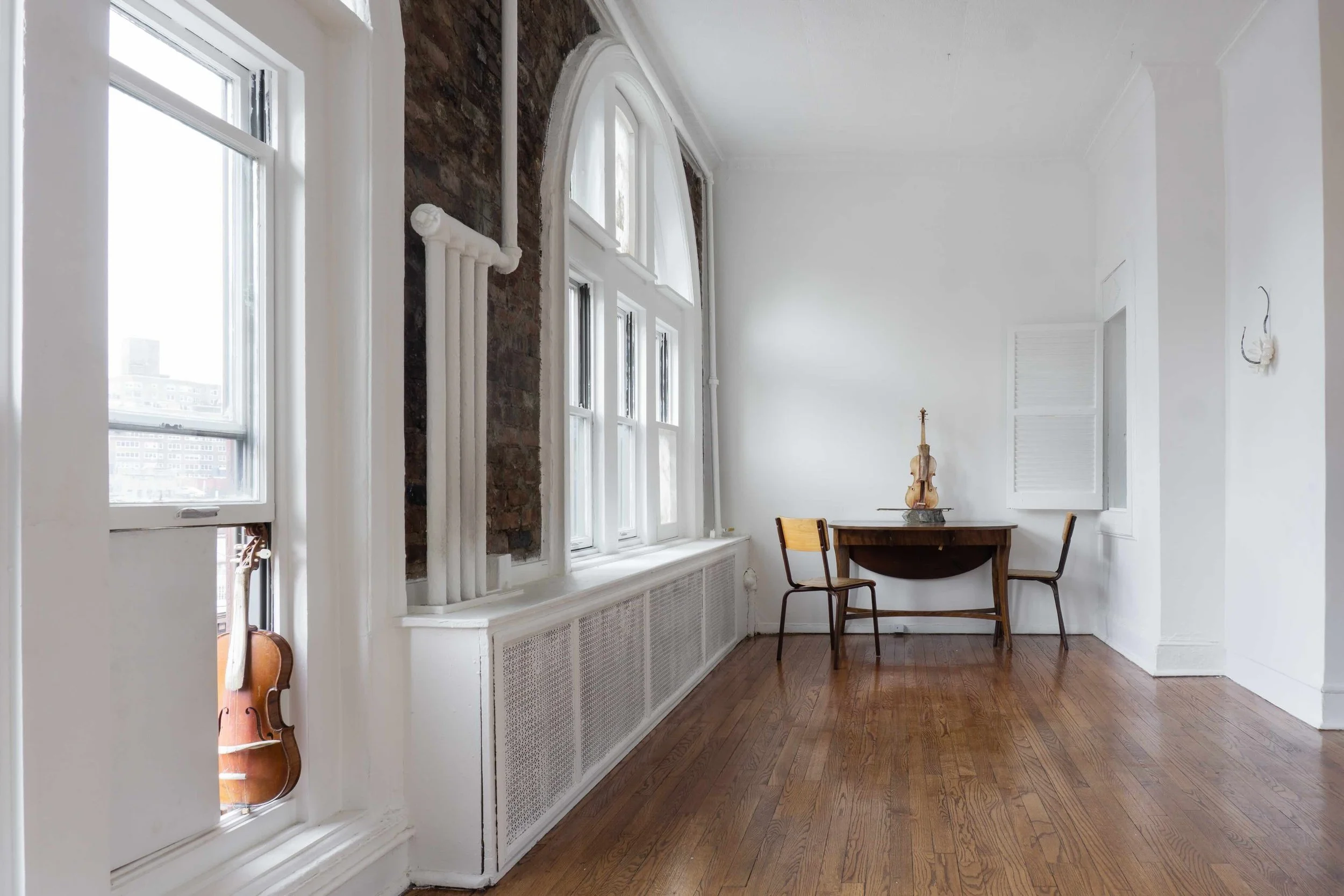 An apartment with white painted walls, exposed brick, and arched windows features sculptural works, including a violin-like object propped in a windowsill and a table with two chairs supporting another violin-like work.