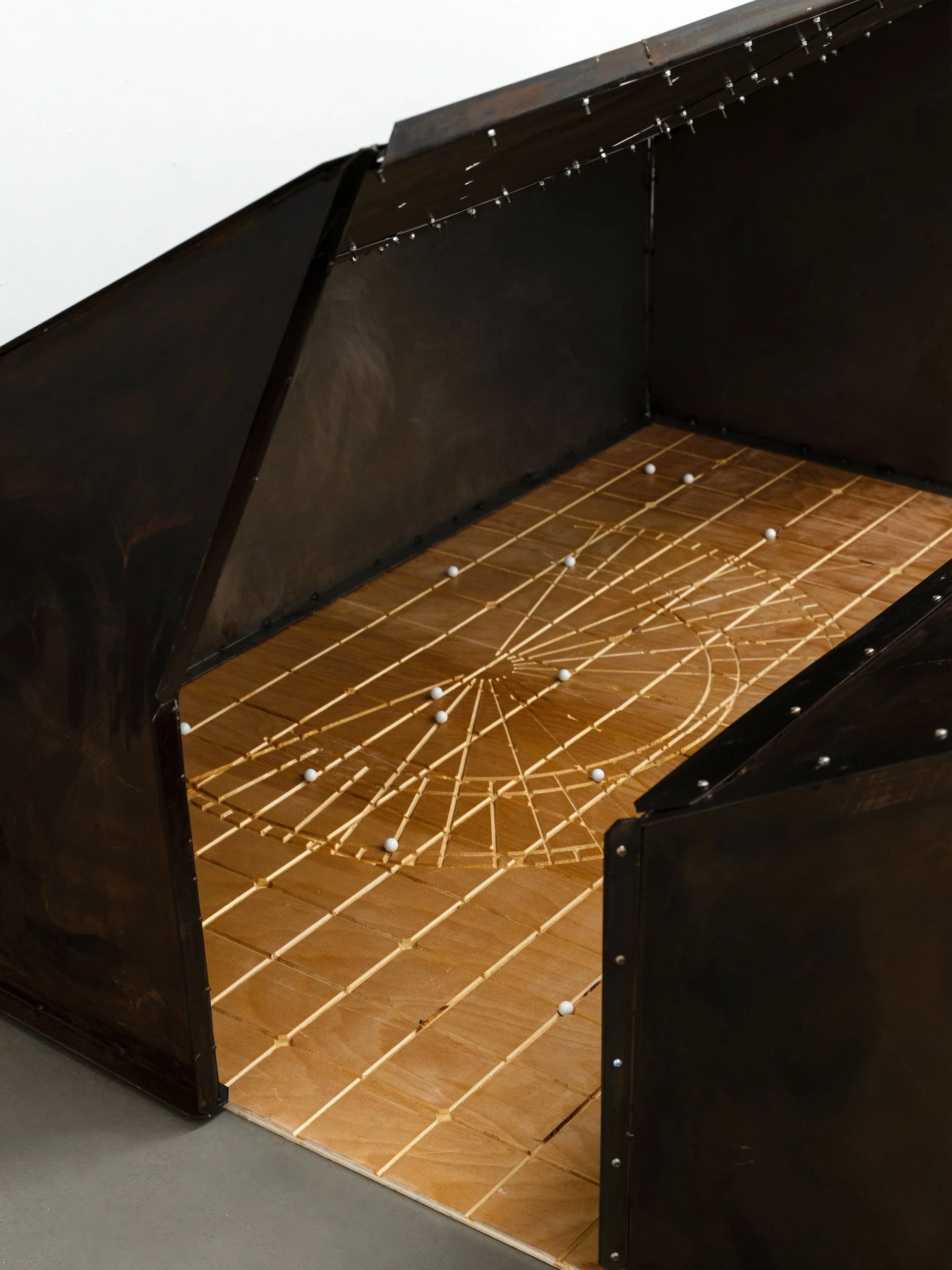 Rusted steel enclosure with an open side, revealing a wooden floor etched with a circular grid and scattered white balls.
