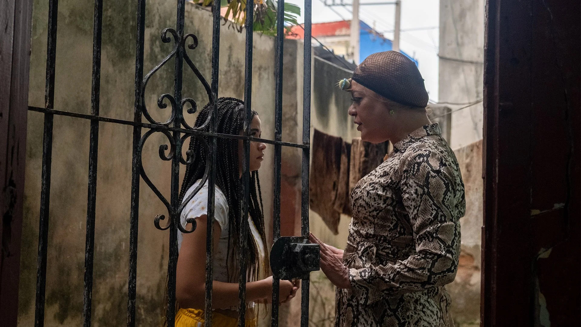 Behind a wrought iron gate door held open by the woman on the left, two women face each other in conversation. They stand in the midst of run-down concrete buildings.