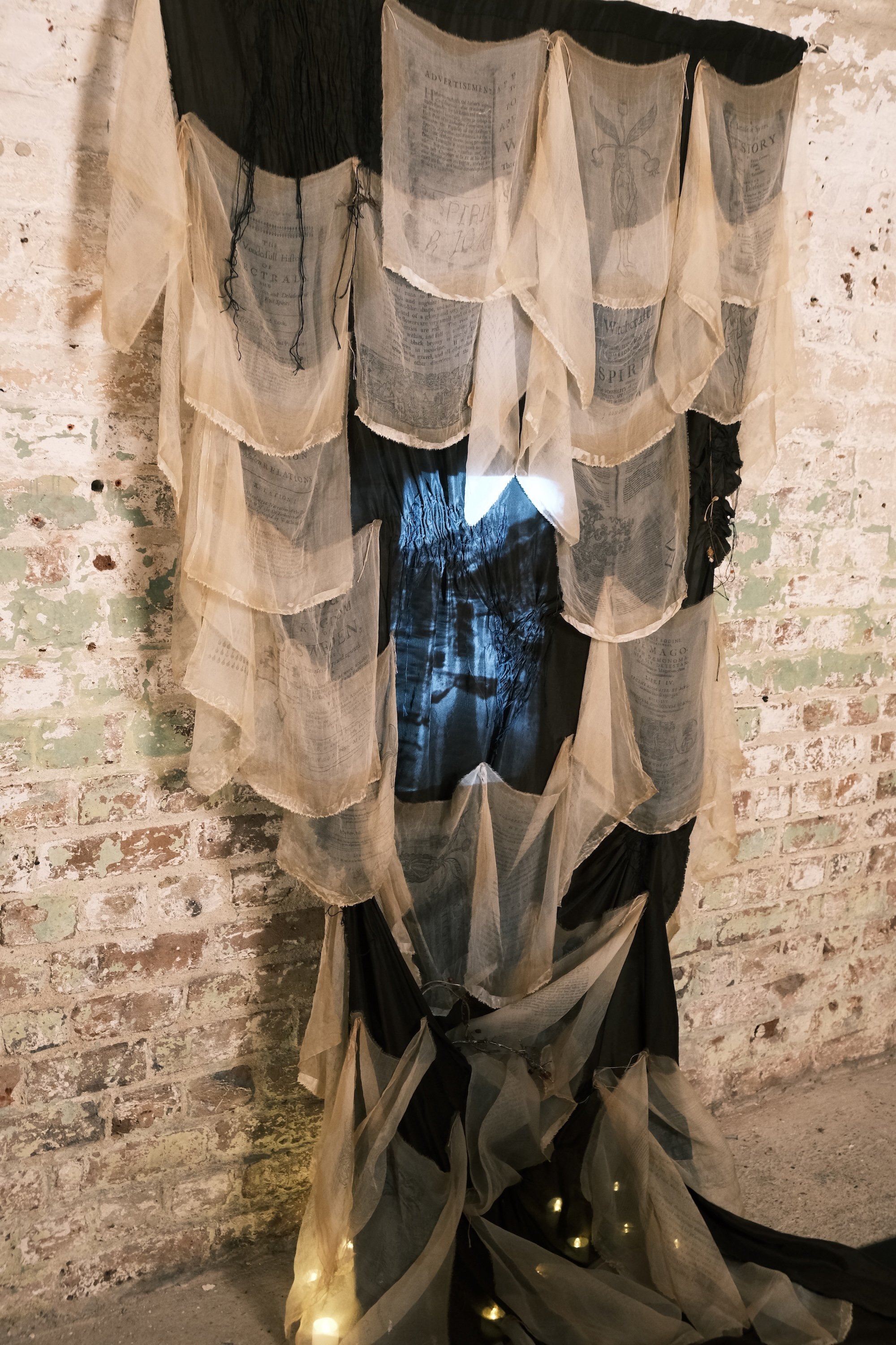 Sheer fabric panels printed with archival text drape over a dark backing, forming a translucent, layered textile installation against a brick wall.