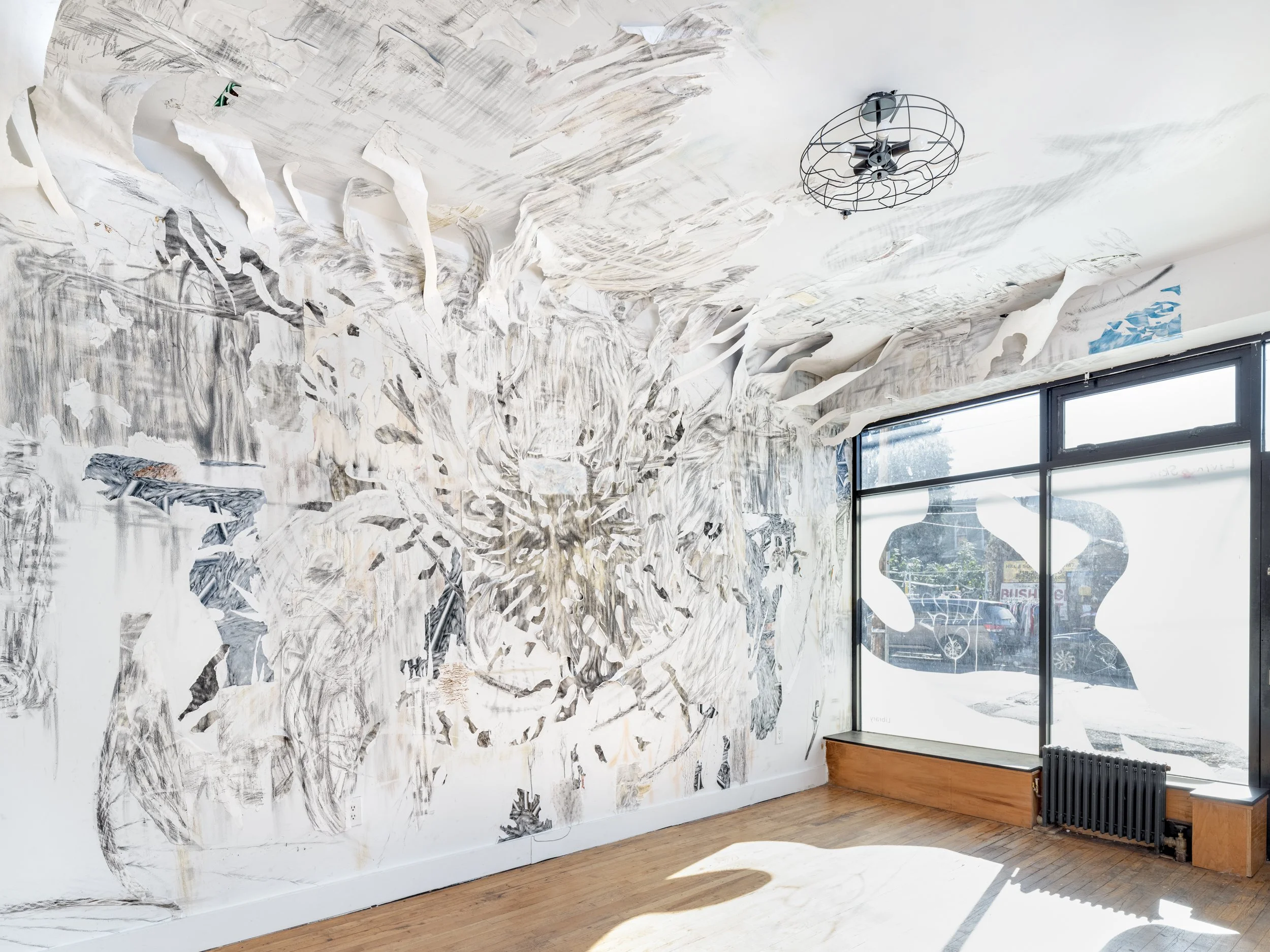 A brightly-lit room with large, street-facing windows has white walls decorated with graphite markings and strips of wallpaper-like shreds attaching to wall and ceiling.