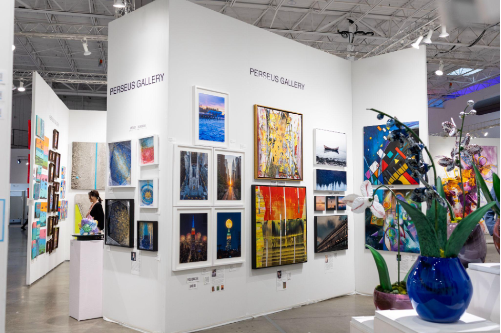 A wide shot of a brightly lit Perseus Gallery art booth, showcasing a large collection of framed photographs, colorful abstract art, and a blue glass vase with flowers in the foreground.