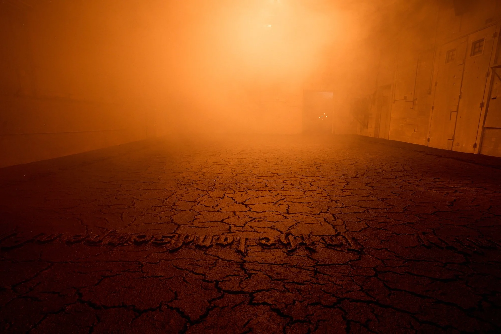 Large indoor installation of cracked, earth-like ground under orange haze, with faint text inscribed across the surface.