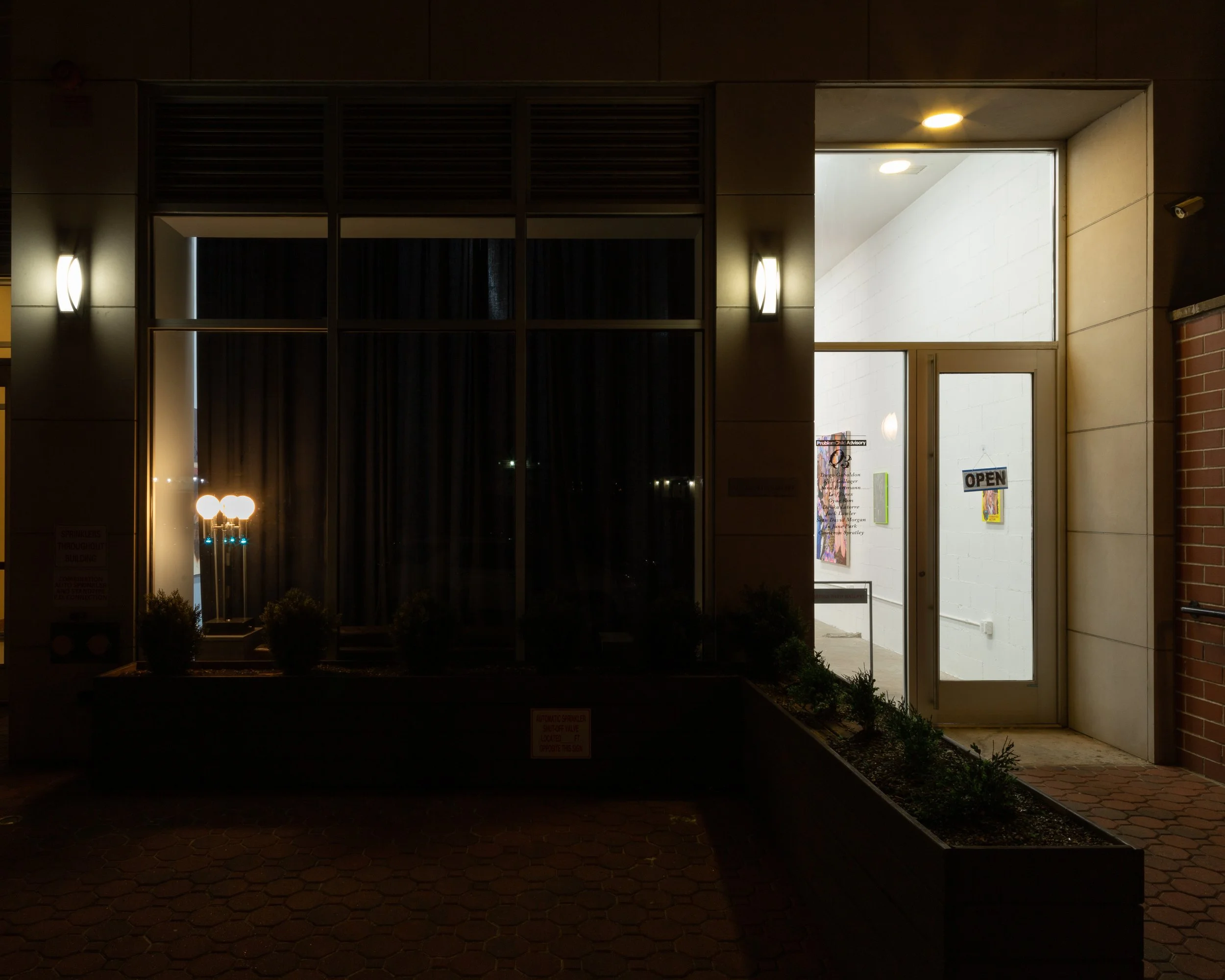At nighttime, the facade of a building front is dimly lit by sconces and lamps. The main windows are covered by dark curtains. Through the glass door, an "open" sign hangs, and brightly lit works on white walls can be seen within.
