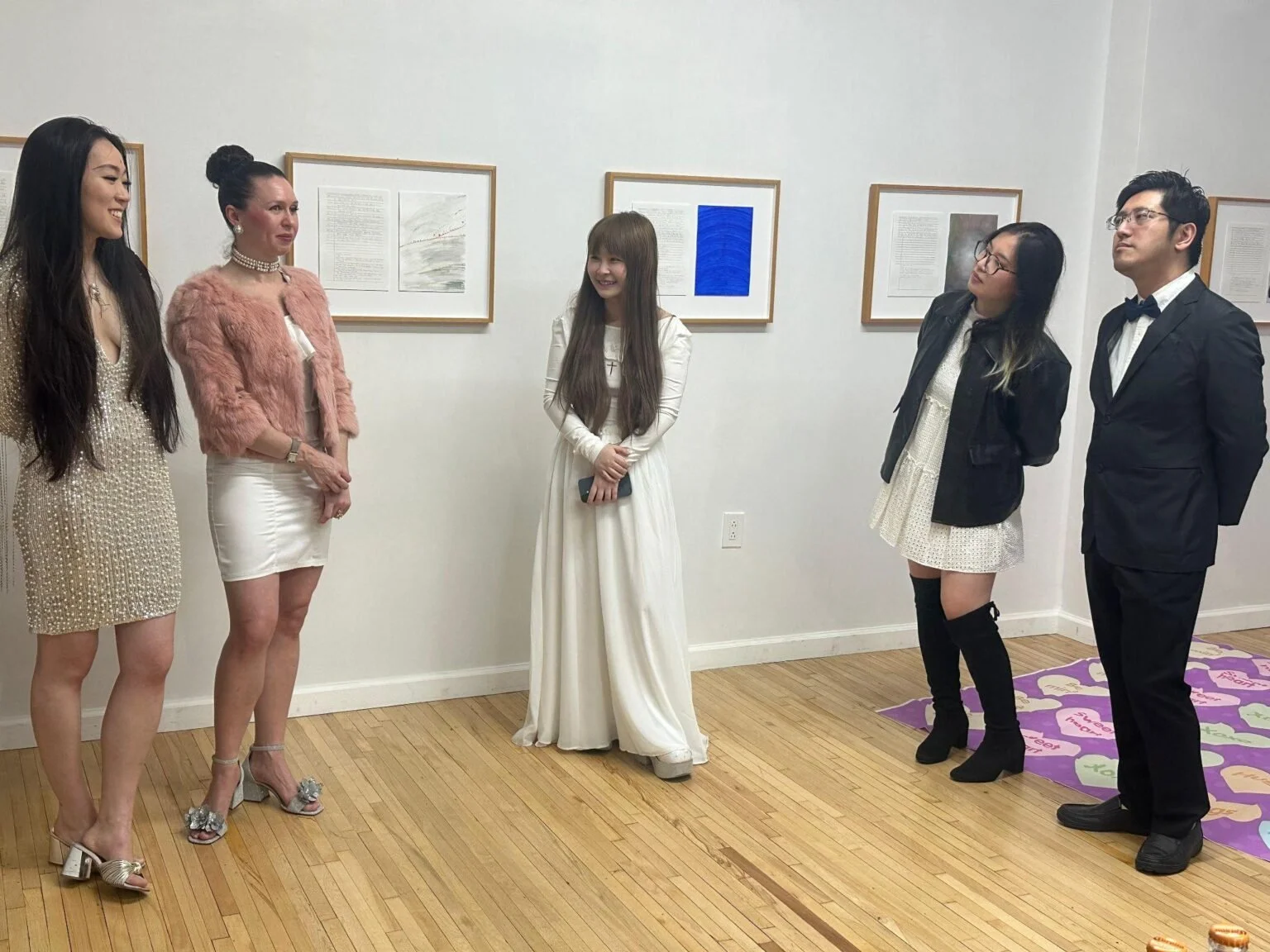 A group of five people dressed in formal and semi-formal attire stand in a white-walled gallery, conversing near framed artwork.
