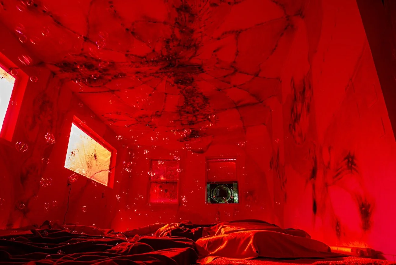 A room lit in red light features patchwork designs on the ceiling and walls. A mattress and sheets lay haphazardly on the floor. Bubbles float through the room.