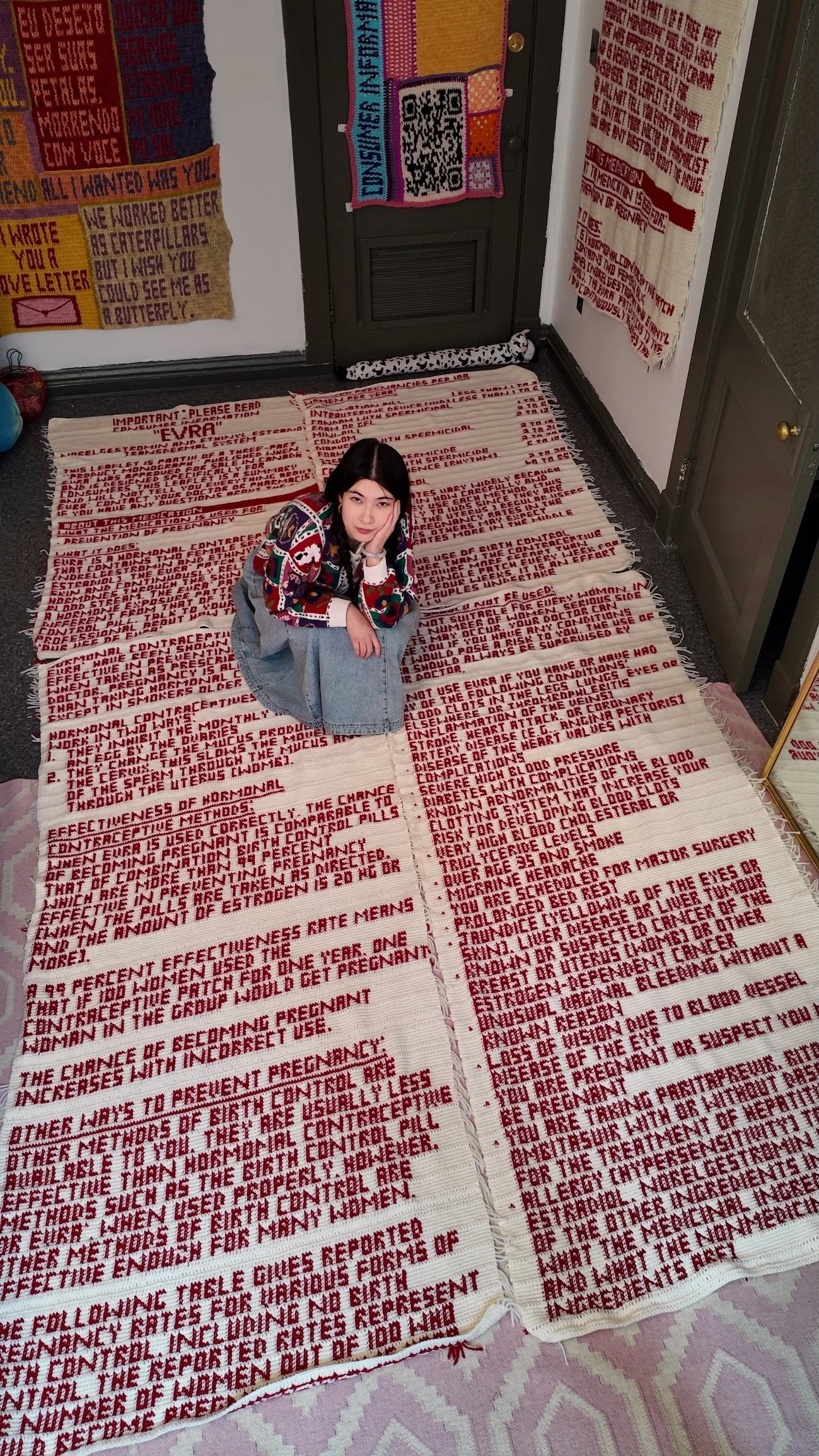Artist seated beside a large crocheted textile densely stitched with red text, displayed on the floor in a gallery corner.