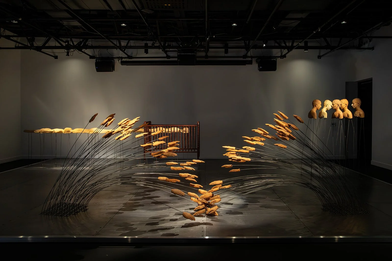 A dimly-lit room hosts multiple sculptural artworks: in the center, flexible metal rods affixed with clay hands stem from two places in the floor and meet in the middle, bending into each other like sheafs of wheat.