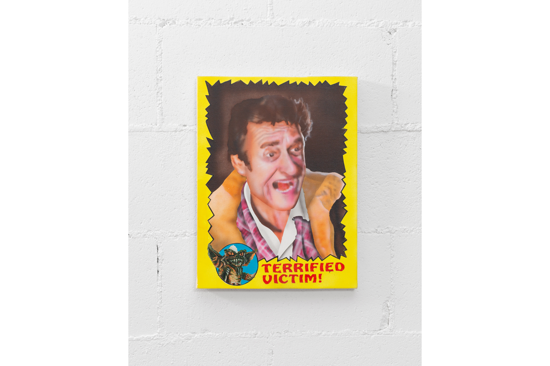 Mounted to a white wall, a graphic work features an airbrushed man with an open-mouthed expression. The border is solid yellow with an irregularly spiked interior. At the bottom, red text reads "terrified victim!" with a gremlin illustration.