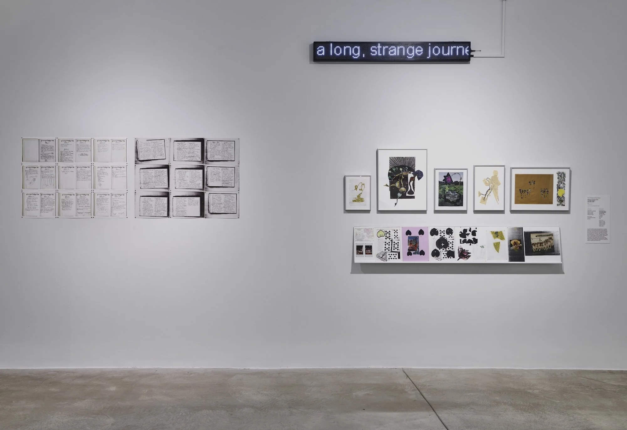 A white gallery wall displaying handwritten text panels, small framed mixed-media works on a shelf, and an LED sign reading “a long, strange journ—”.