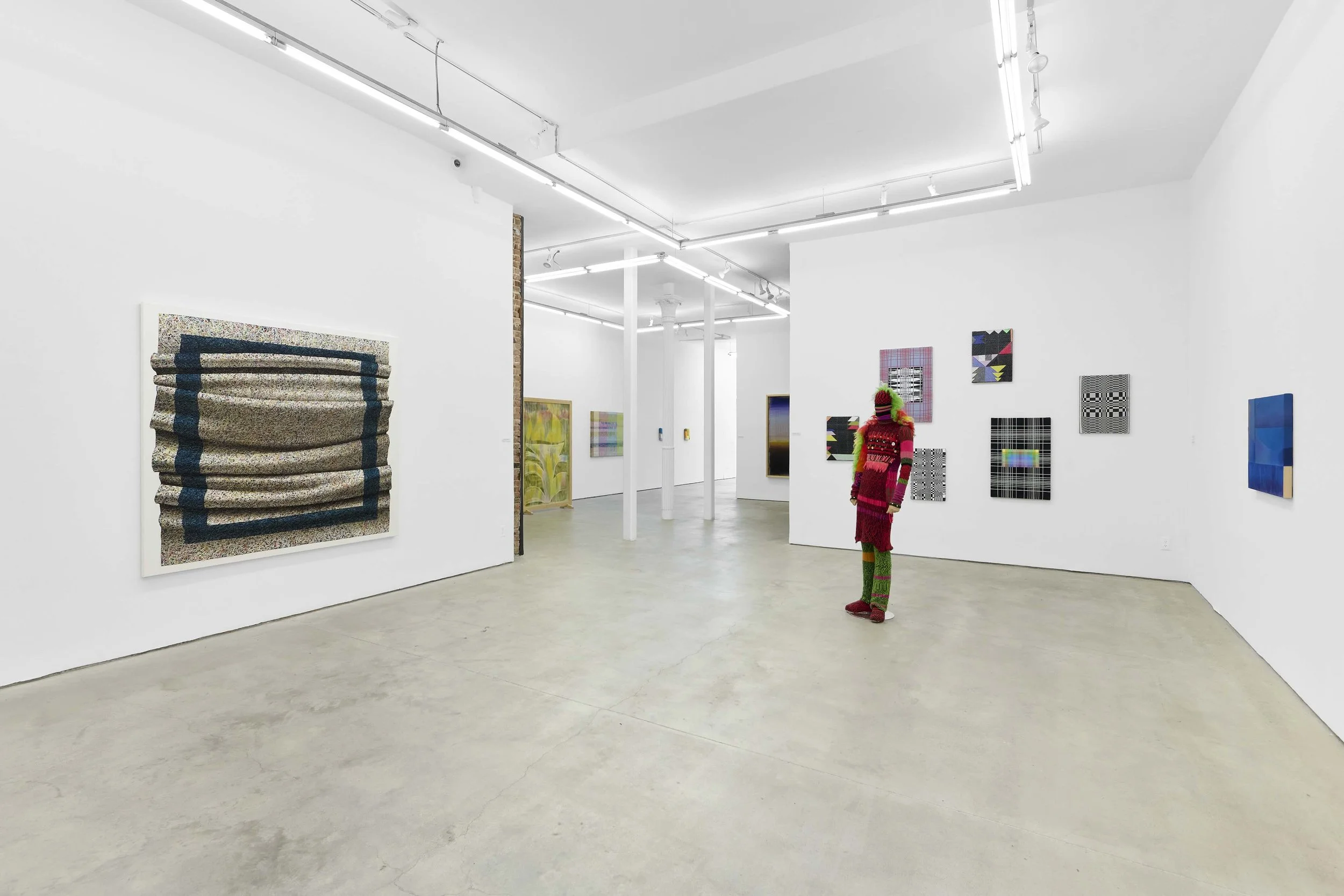 A white gallery complex features multiple textile and wall works of various sizes and colors. In the floor space, a colorful fabric mannequin, dressed in ornate designs, stands.