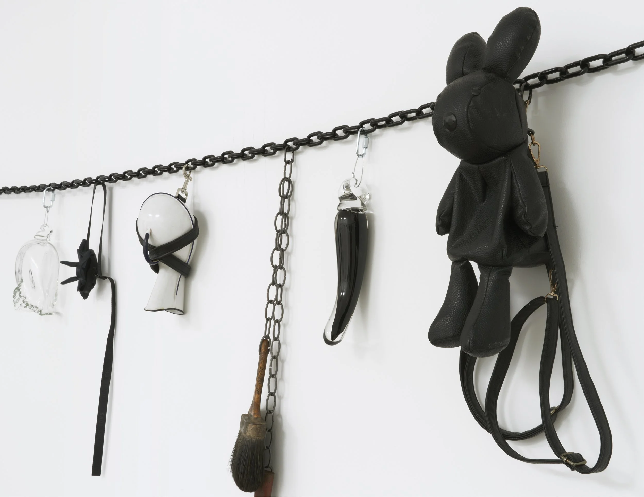 A black chain strung against a white wall supports a variety of clipped-on attachments. The attachment objects include a black leather bunny, black straps, and a glass phallus.