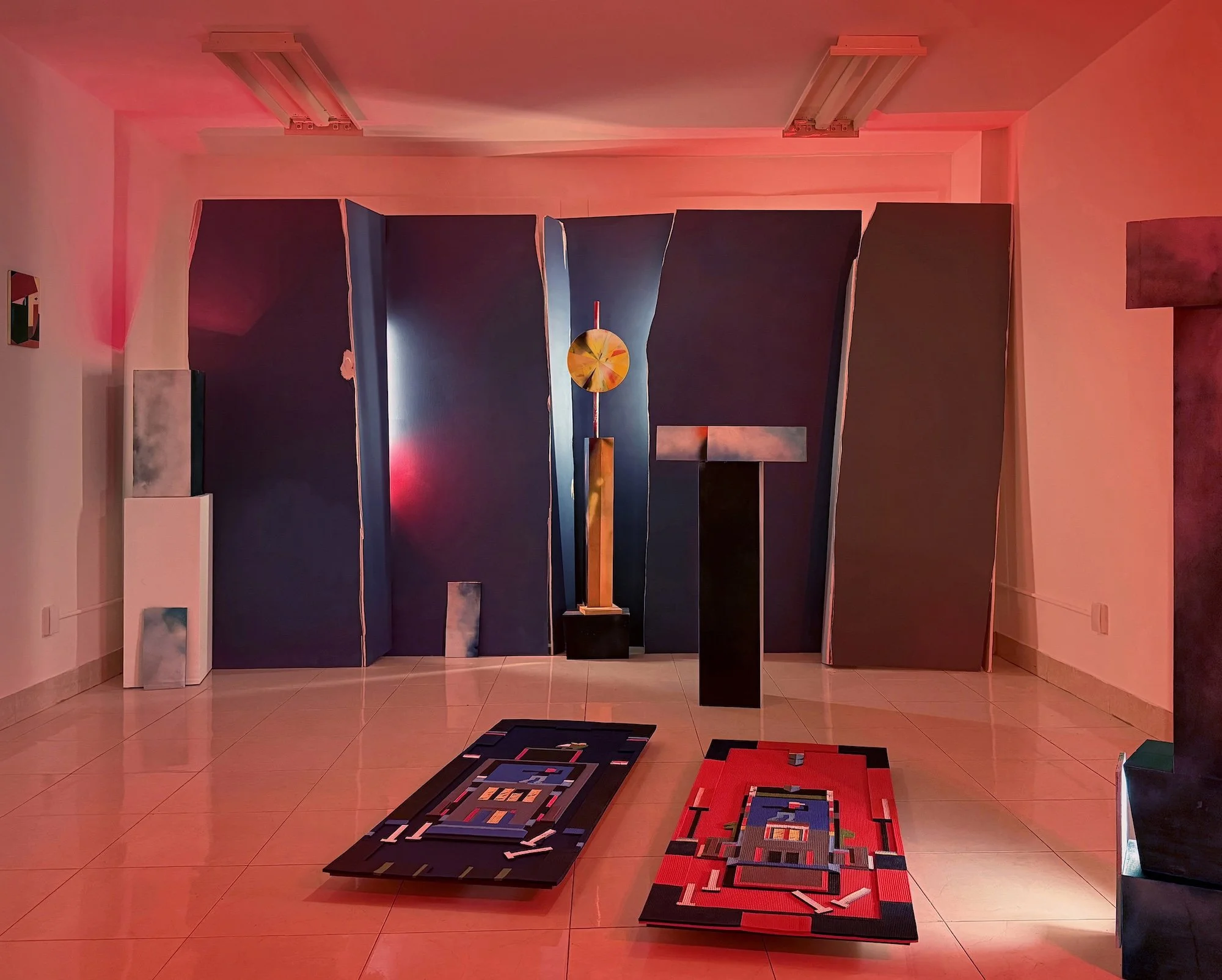 Wide installation view showing tall sculptural panels, columnar forms, and two rectangular textile works laid on the gallery floor under red lighting.