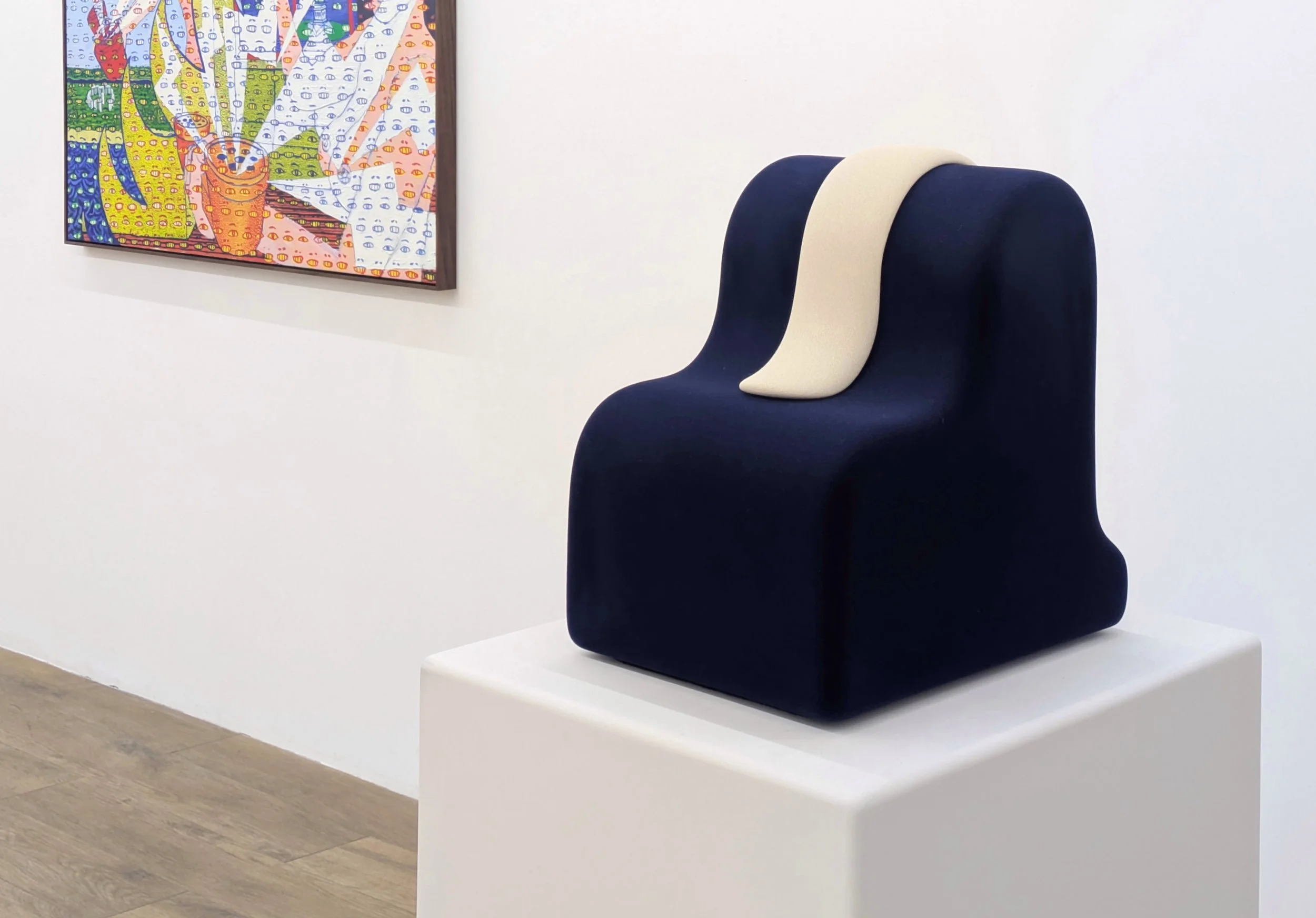 A white gallery room features a colorful painting on its wall and a white plinth supporting a smooth, chair-like sculpture finished with a dark blue, velvet-like material. It features one central raised white stripe finished in the same material.