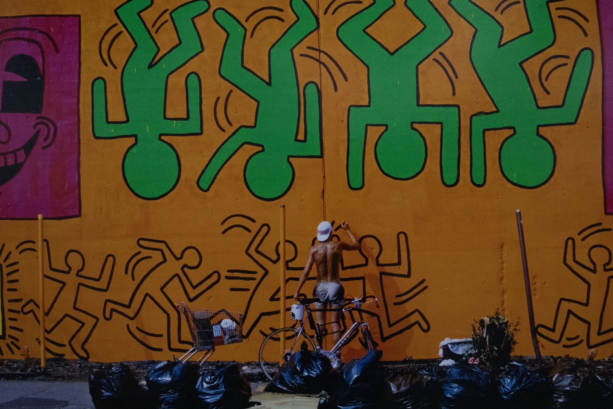 Shirtless artist paints green, dancing figures on a large yellow wall mural at night.