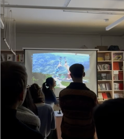 A person stands in front of a projector screen displaying a bright blue landscape, presented to an audience in a room with bookshelves.