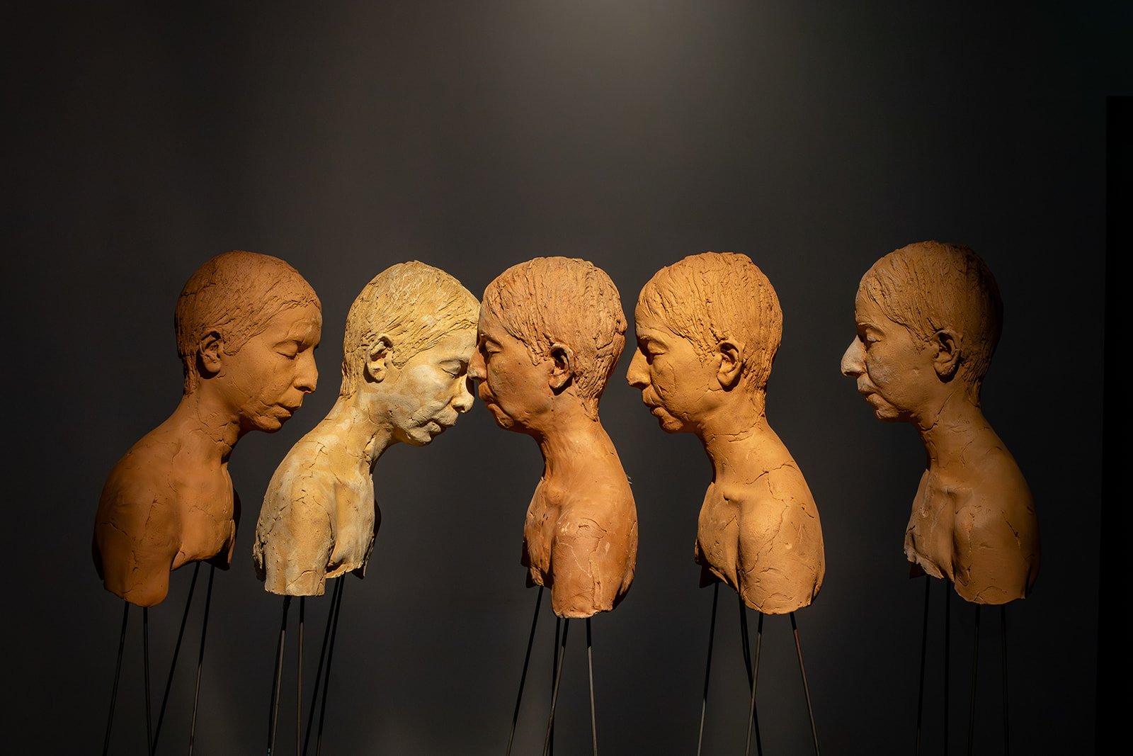 In a darkly-lit gallery room, a sculpture stands, comprised of five busts affixed to the top of standing, flexible metal rods. The busts resemble figures with short hair and their eyes closed. Three of the busts face left and two face right.