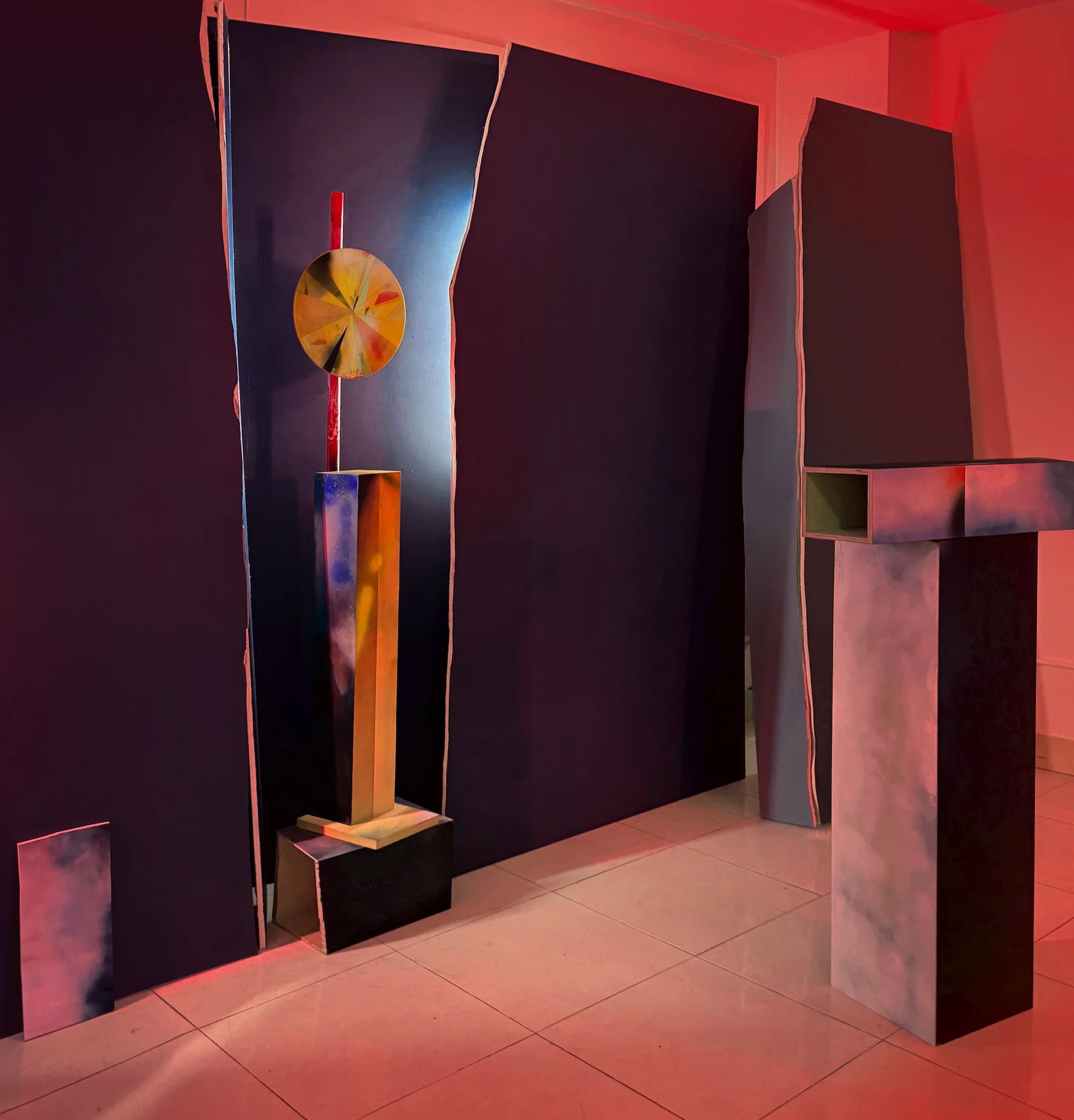 Installation view with freestanding sculptural columns and wall panels, lit in red and blue tones, with a circular geometric element mounted vertically.