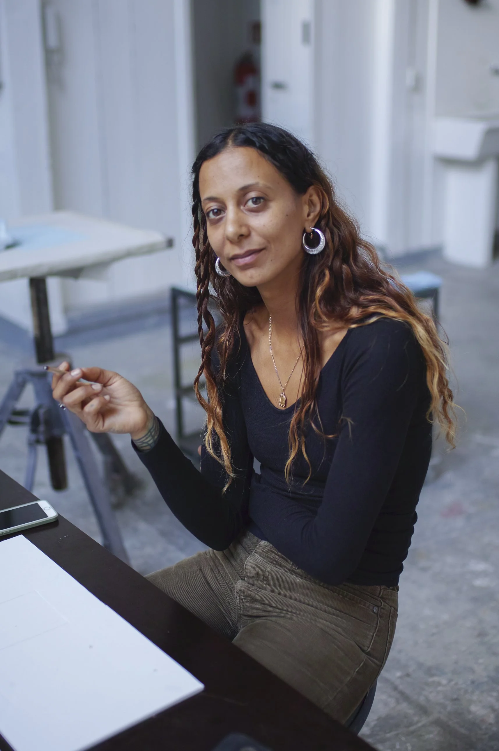 A person in a black tip and grey jeans with long, wavy brown hair with blonde ends sits at a table and faces the camera, smiling.