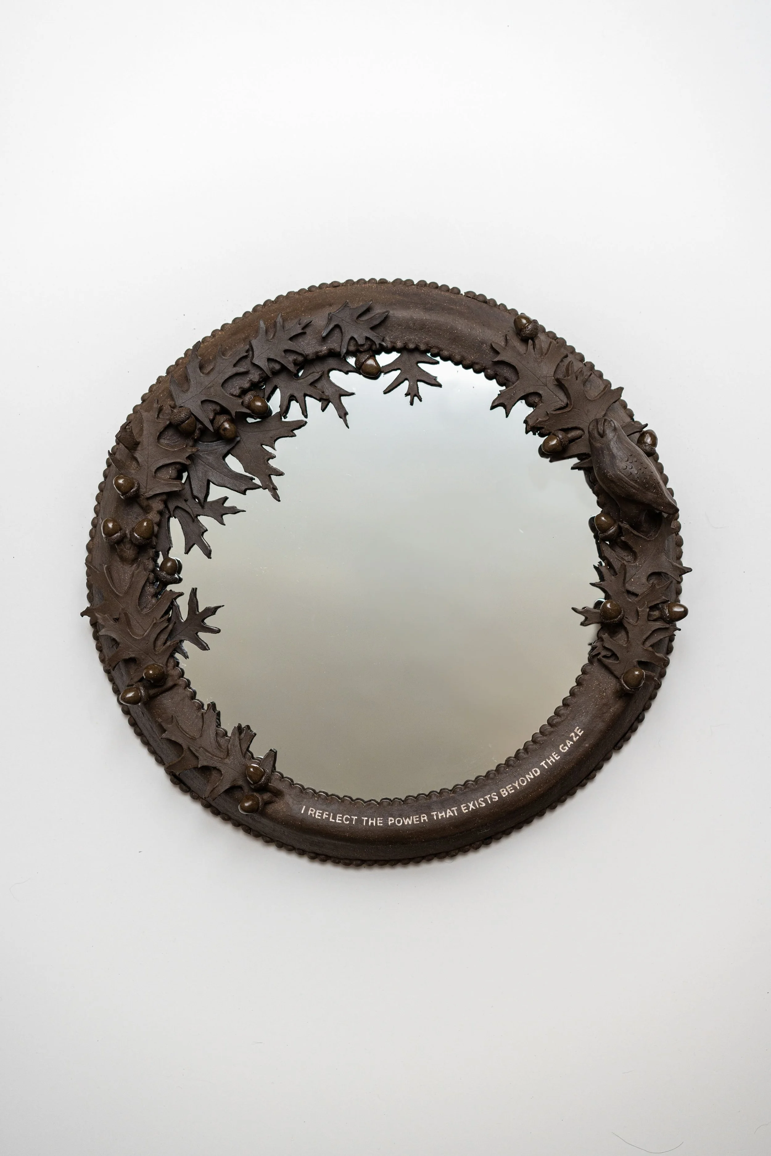 A wall-mounted circular mirror is framed by a thick, brown frame decorated with sculpted leaves, acorns, and a bird from the same frame material. On the bottom, text curved with the mirror reads "I REFLECT THE POWER THAT EXISTS BEYOND THE GAZE."