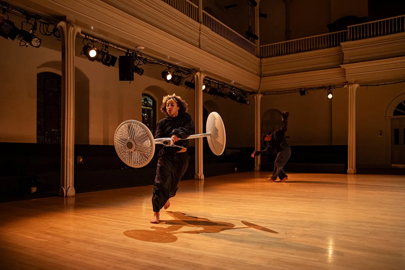 In a balconied performance space, a dancer carries a white standing fan and runs about the space. In the background, another performer gestures with outstretched hands and bent legs.