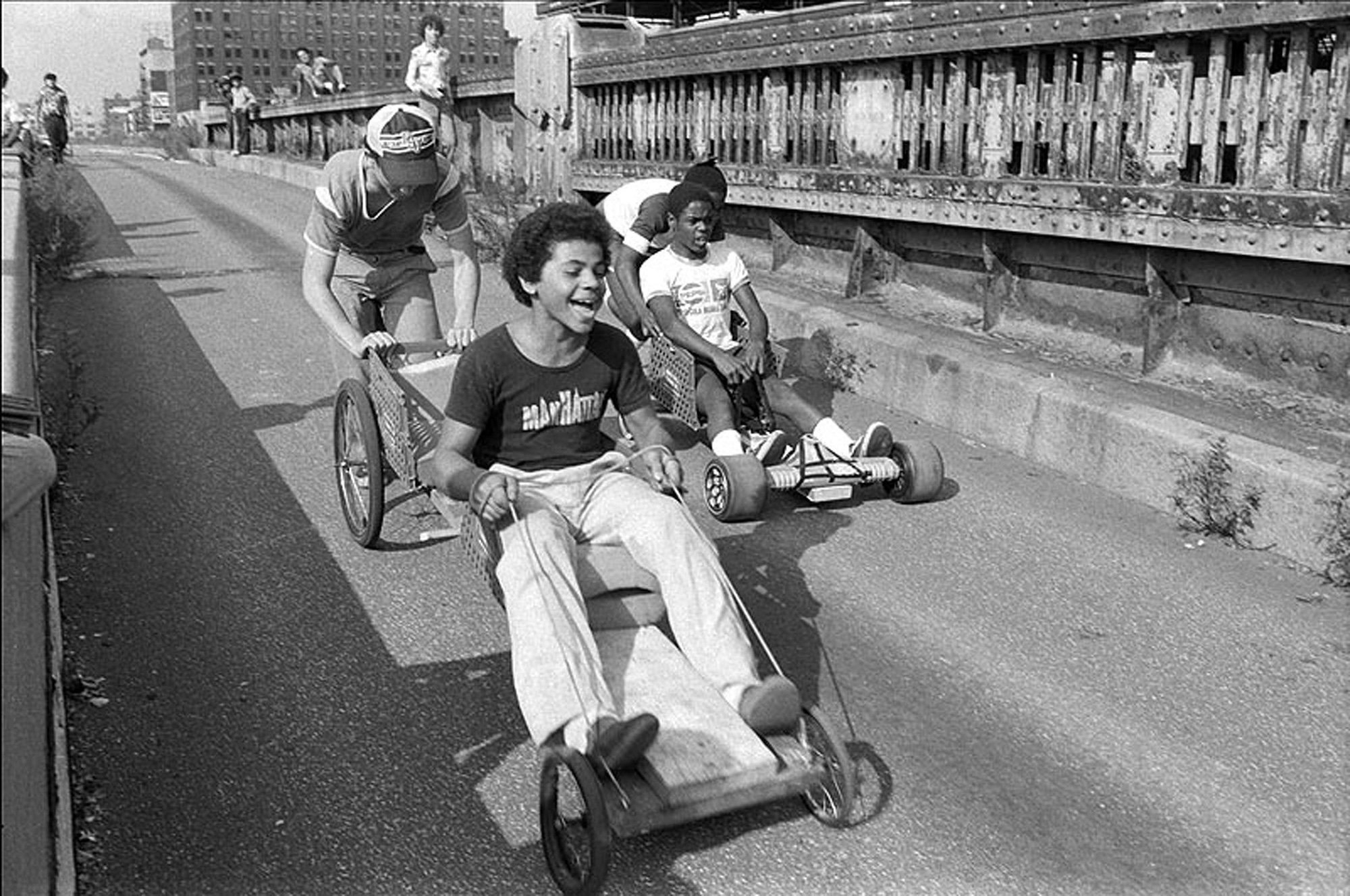 Children riding makeshift carts down a paved path near an industrial bridge.