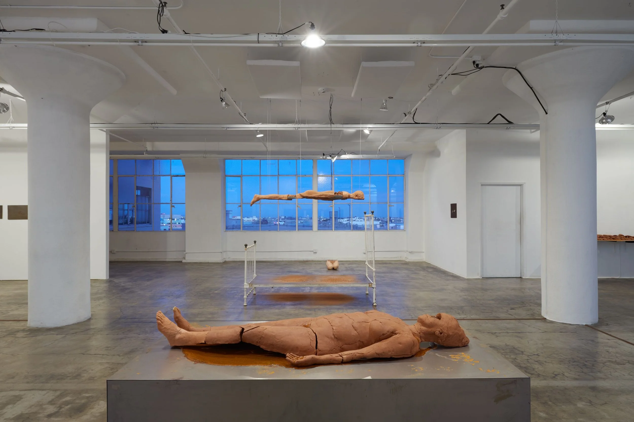 In a large, warehouse-like gallery space, a centrally framed sculpture consisting of a fractured clay body with its face destroyed sits on top of a grey plinth. Behind the sculpture, a bisected clay body hangs facing the floor, its front half absent.