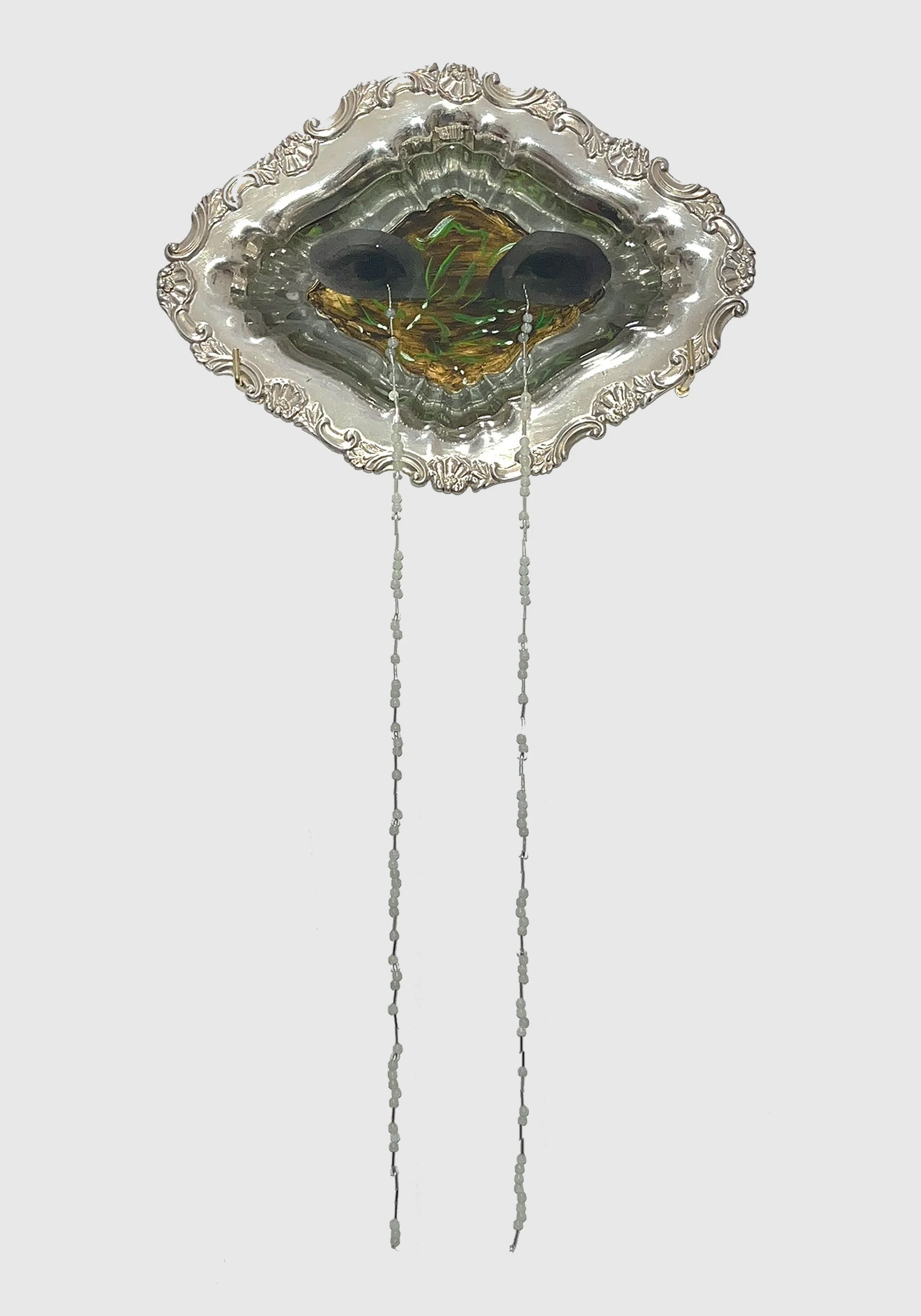 A decorated silver rhombus displays a painted surface within: eyes overlaid on flowering plants in the dirt. From the eyes, two long, thin strings of white beads hang down.